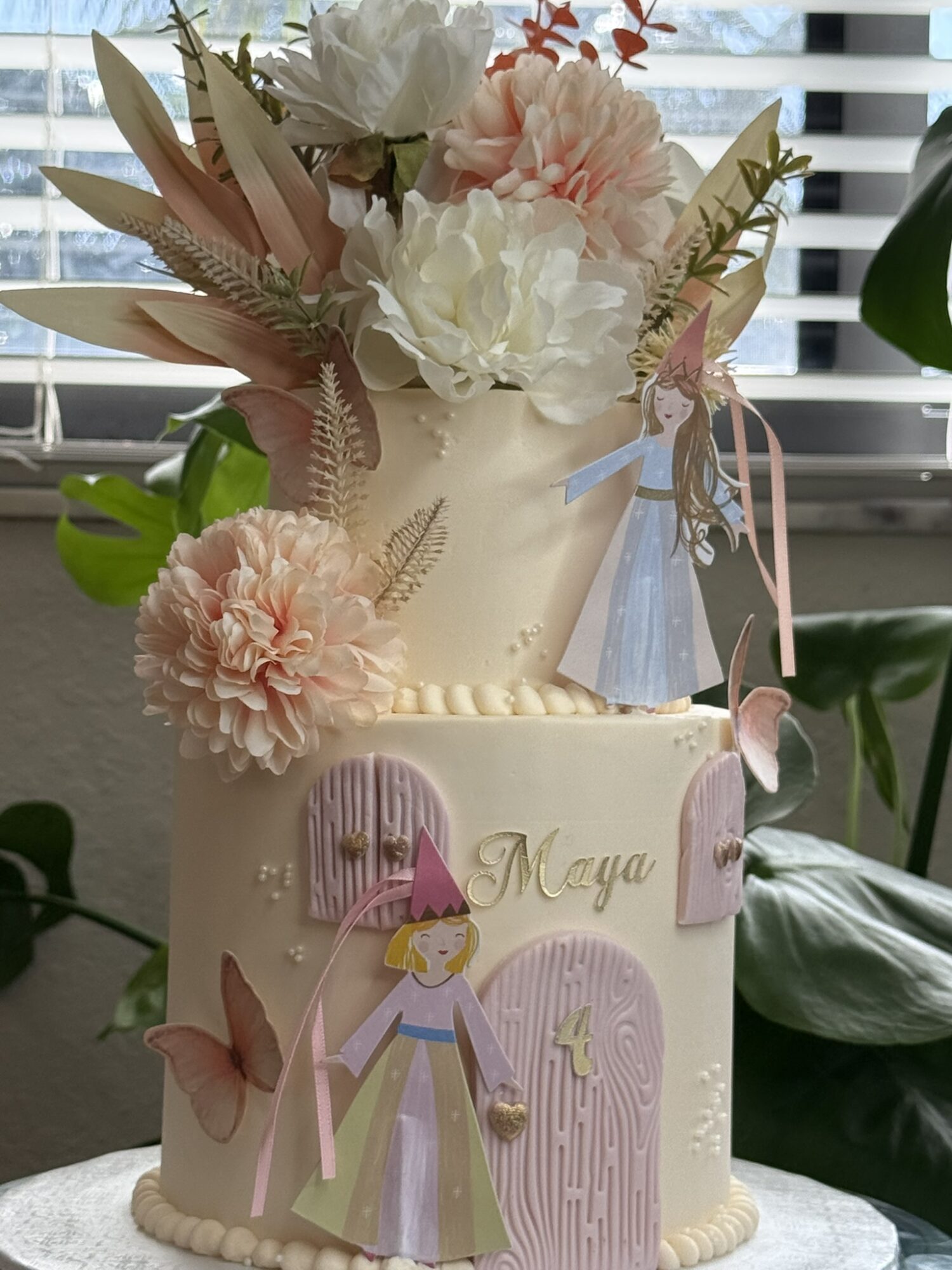 Two-tiered cake decorated with pink and white flowers, a paper princess, and the name 'Maya' for a first birthday celebration.