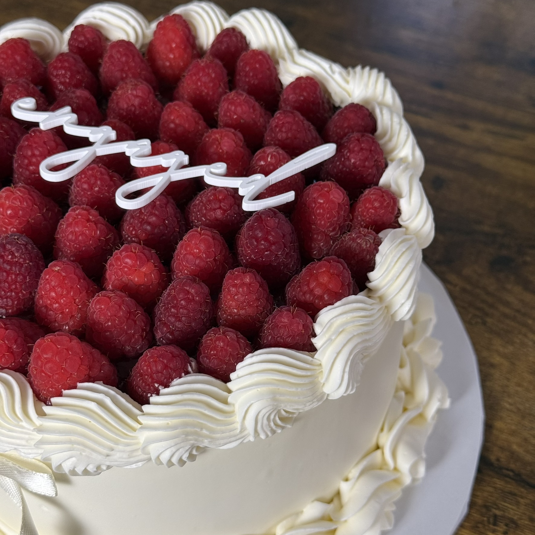 Round cake decorated with raspberries and white frosting, with a white 'Happy' topper, on a wooden surface.