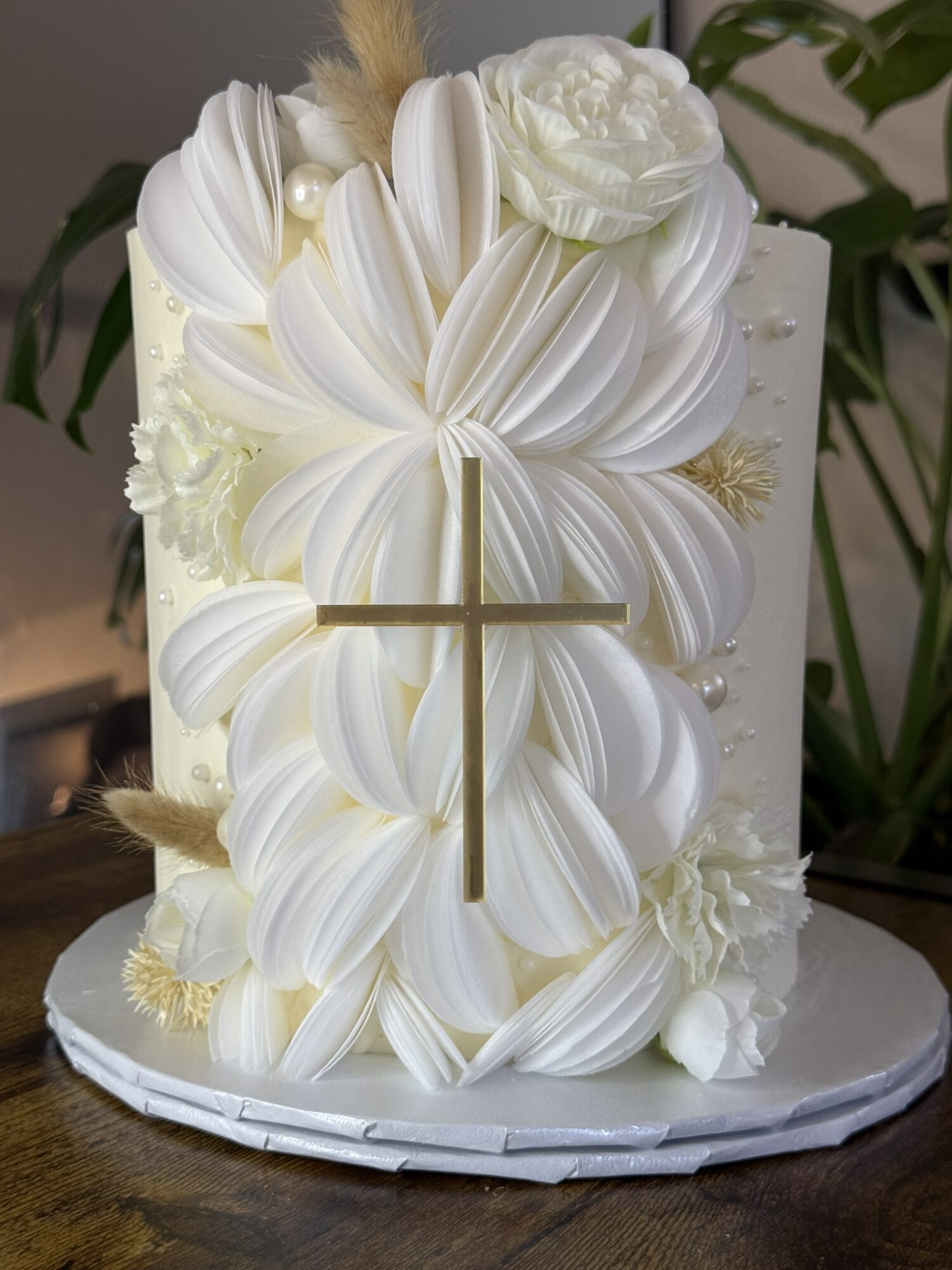 Decorative cake with white floral and ribbon designs, gold cross, and white flower on top, on a white base.