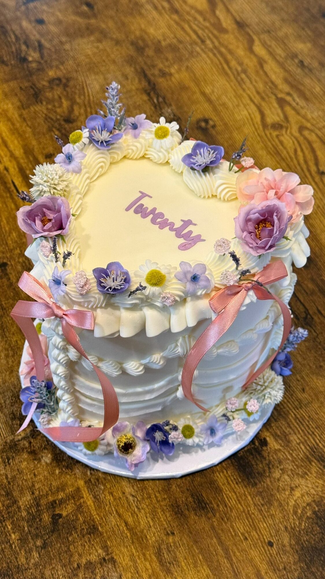 Round decorated cake with purple, pink, and white flowers, pink ribbons, and the word 'Taweta' written on top, on a wooden surface.