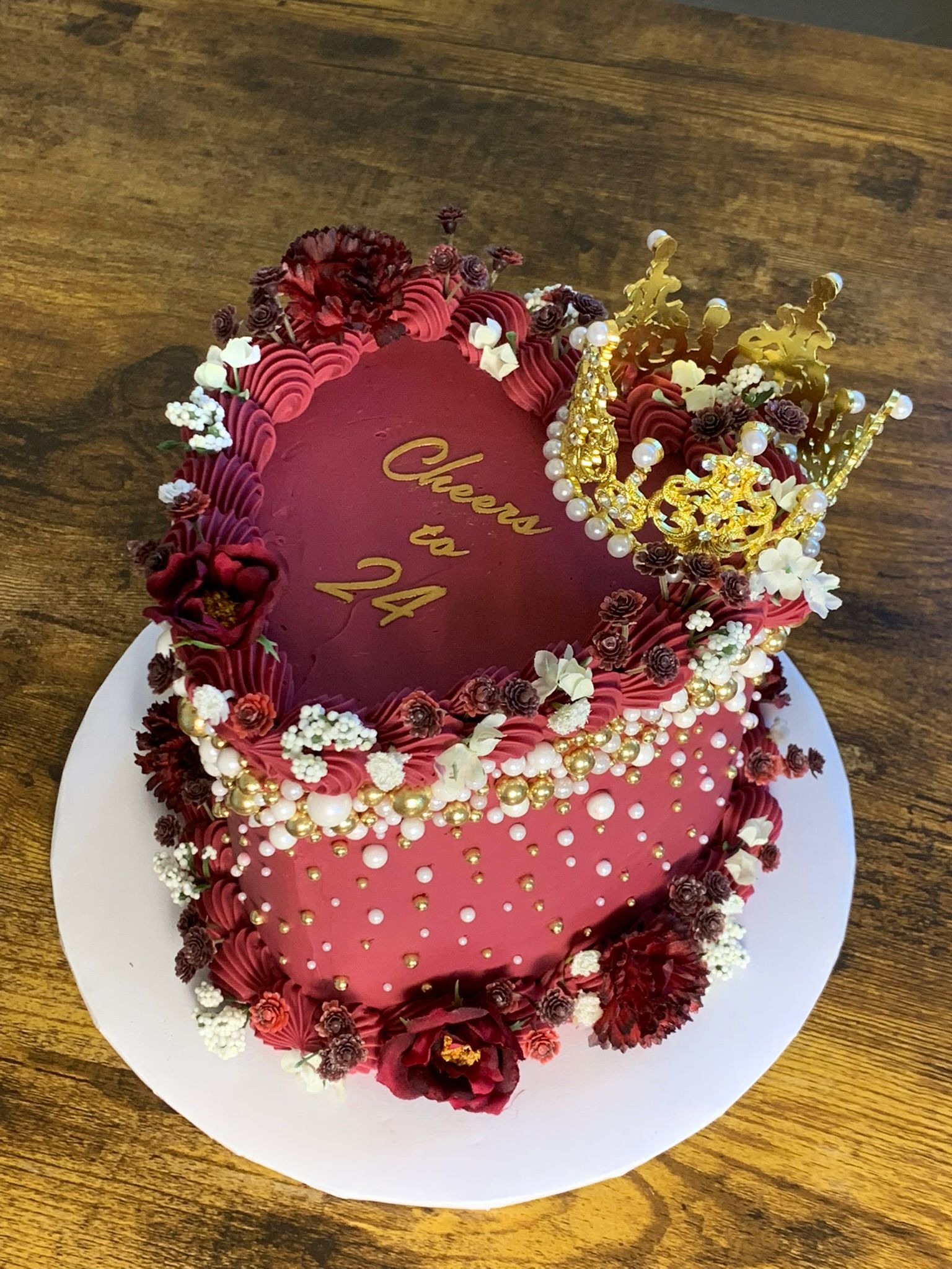 Two-tiered pink cake decorated with red and white flowers, gold beads, and a message in gold script, on a white plate.