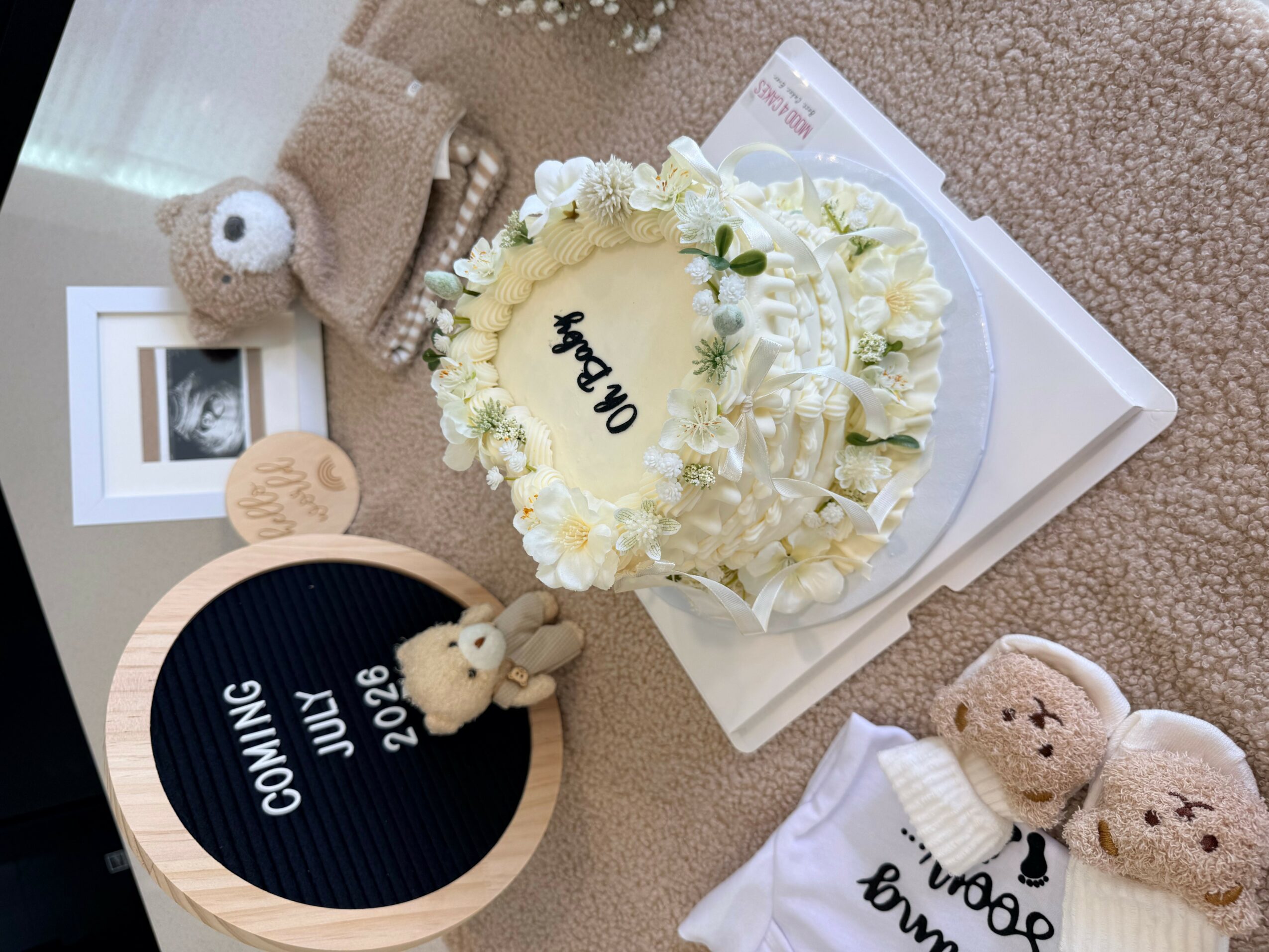 A cake decorated with white flowers and a message, surrounded by plush toys and a round sign with text, on a beige surface.