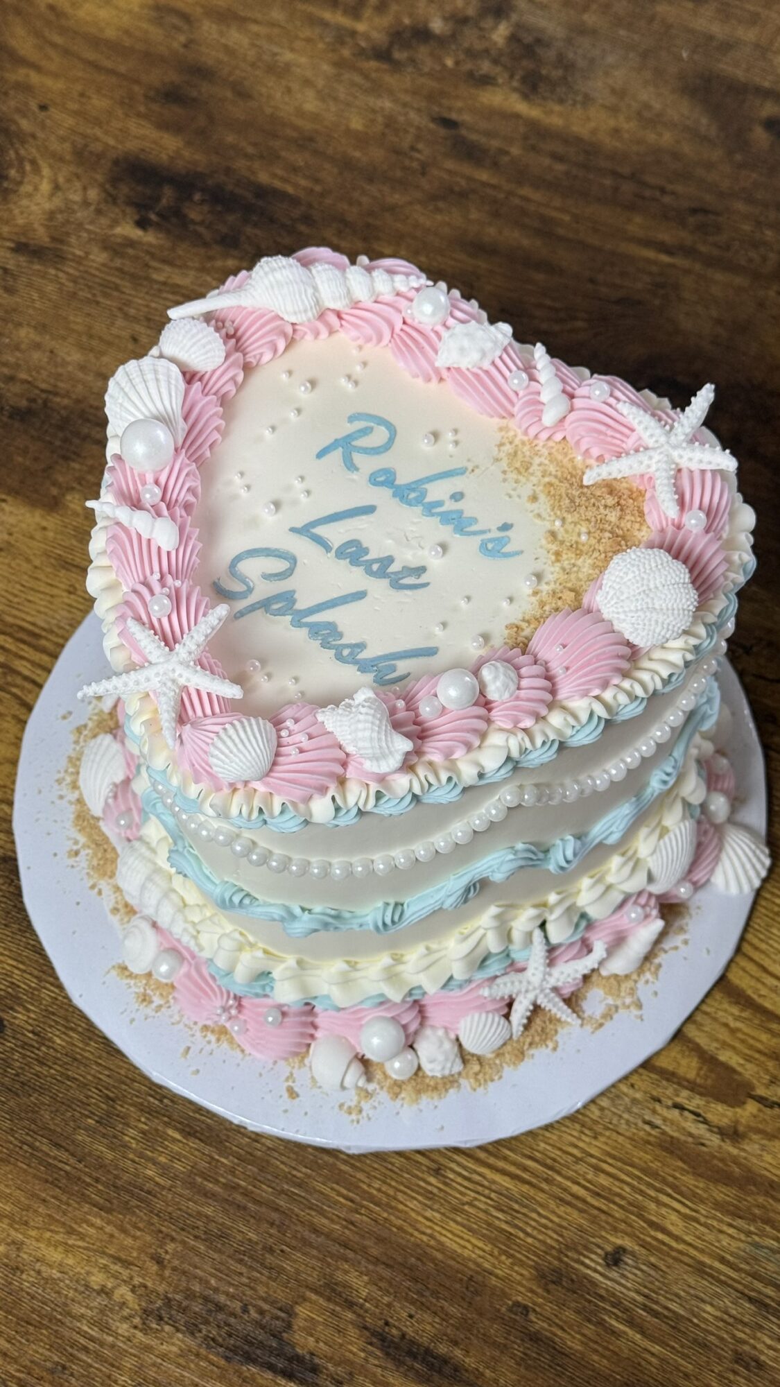 Heart-shaped cake decorated with pink and white icing, shells, and starfish, with a message on top, on a wooden surface.
