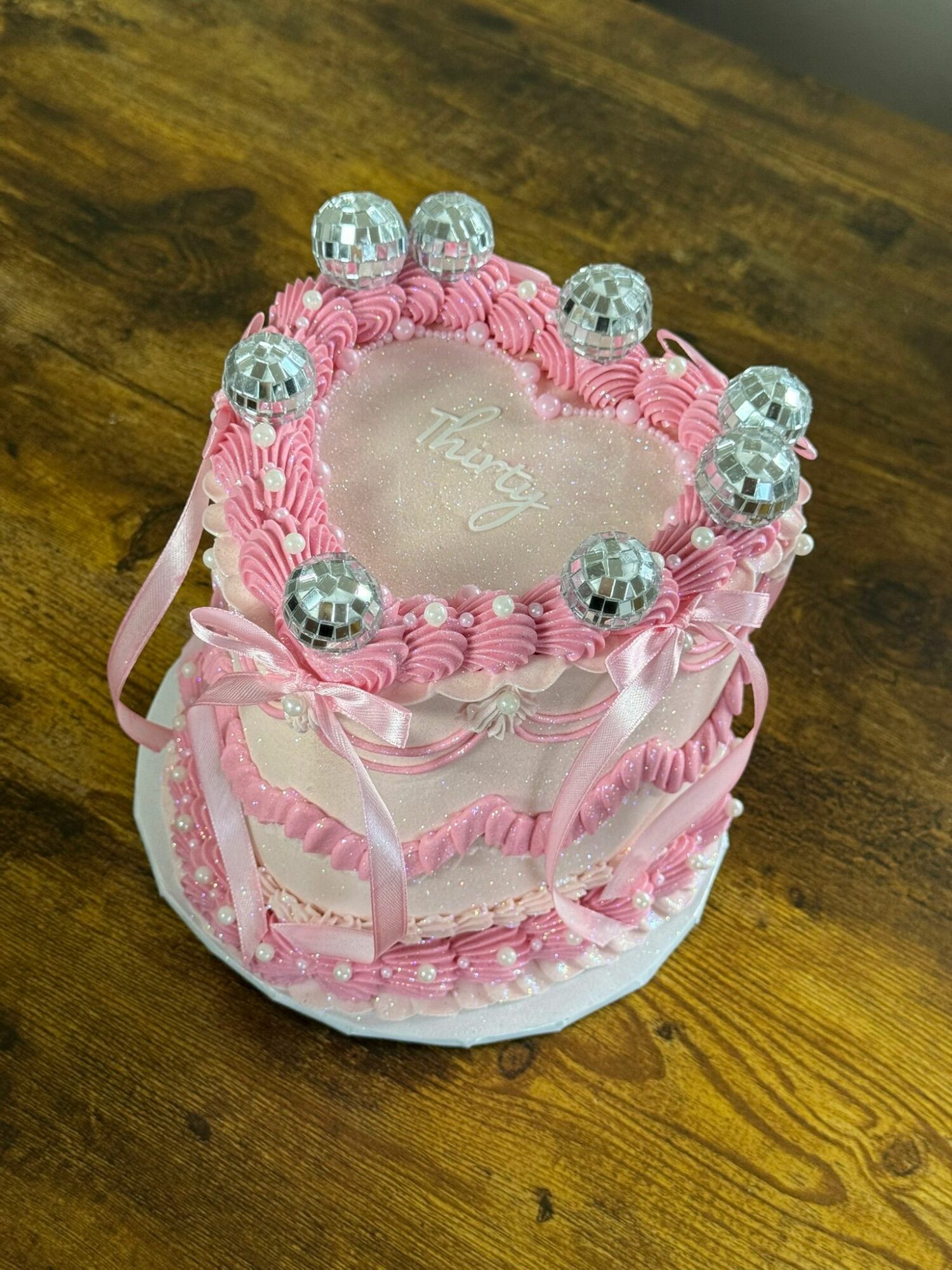 Pink heart-shaped cake with silver balls and decorative pink icing, on a wooden surface.