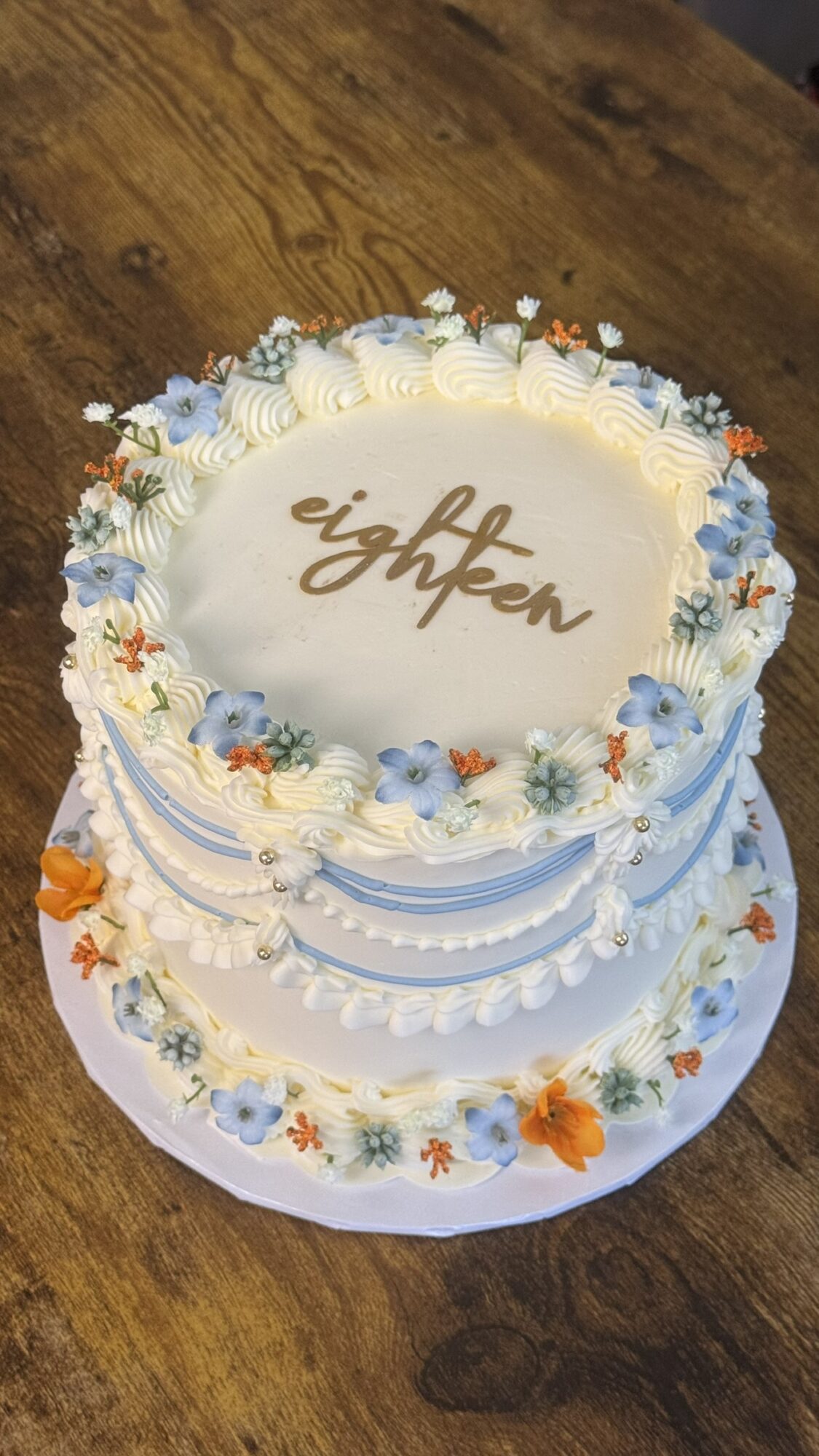 Round white cake with blue and orange floral decorations, and the word 'eighteen' written on top, on a wooden surface.