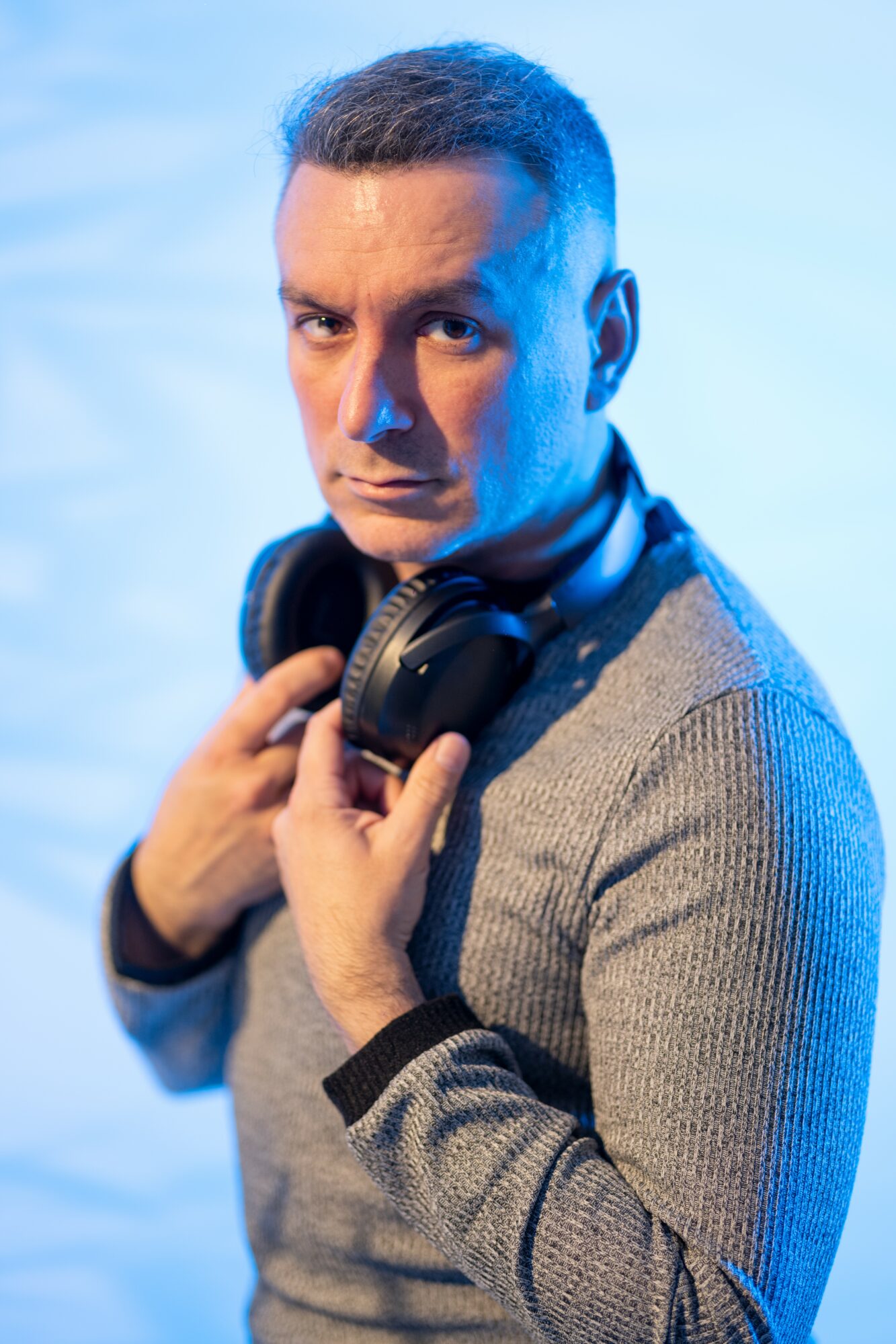 Man with short hair holding headphones around neck, looking at camera, wearing a gray sweater, against a blue background.