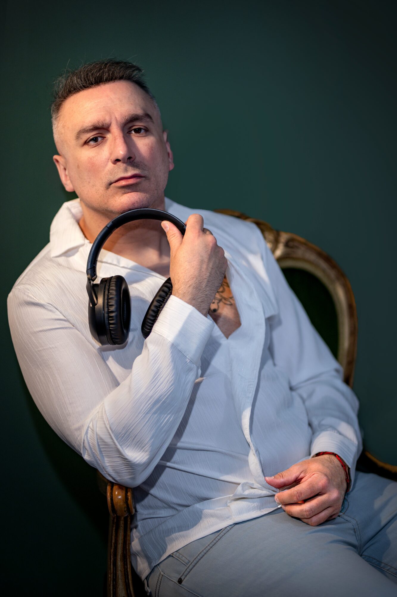 Man with headphones around neck, sitting on a chair, looking at camera, dark green background, wearing white shirt.