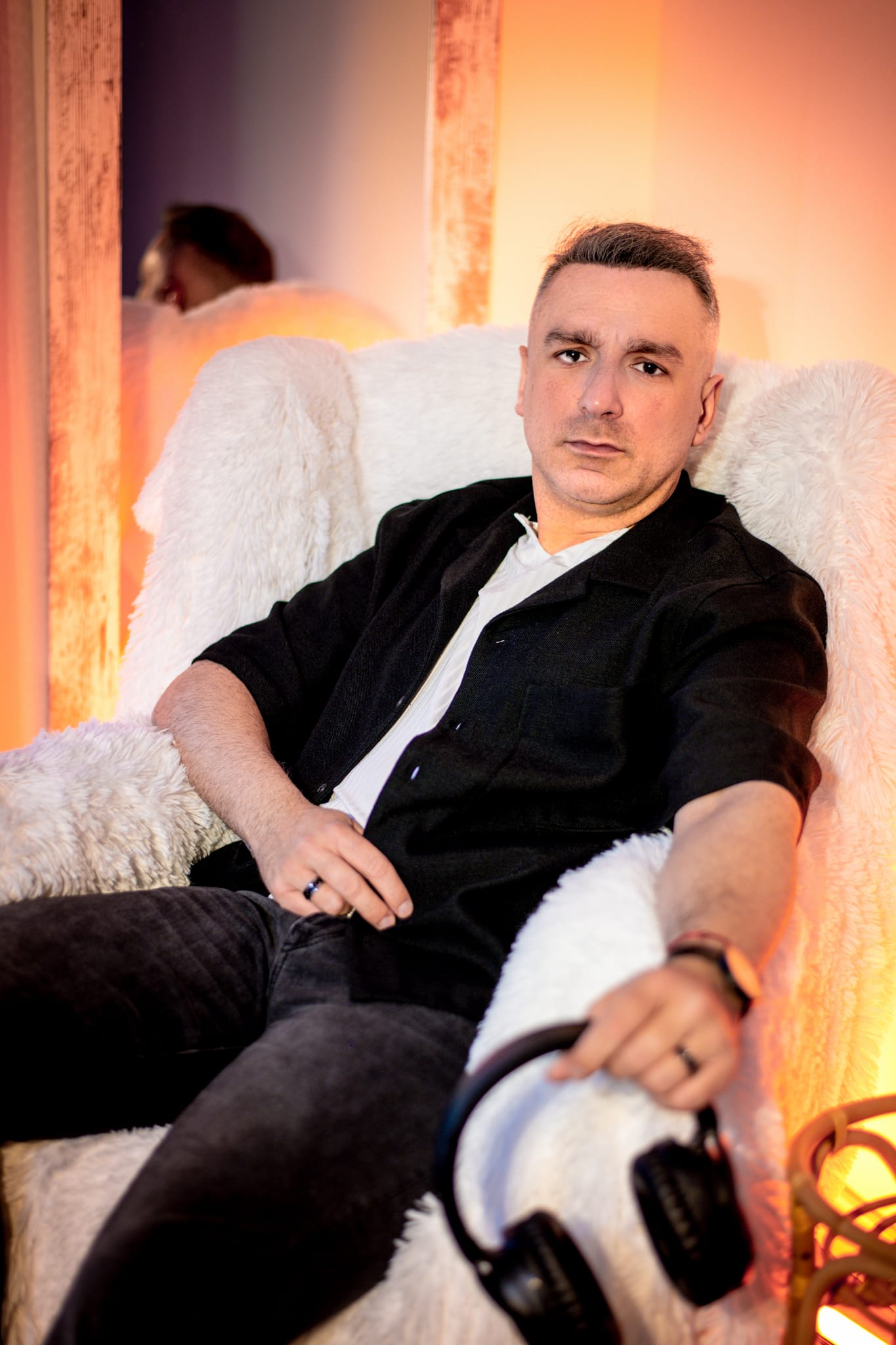 Young man with short hair sitting on a white chair, holding headphones, in a warmly lit room.