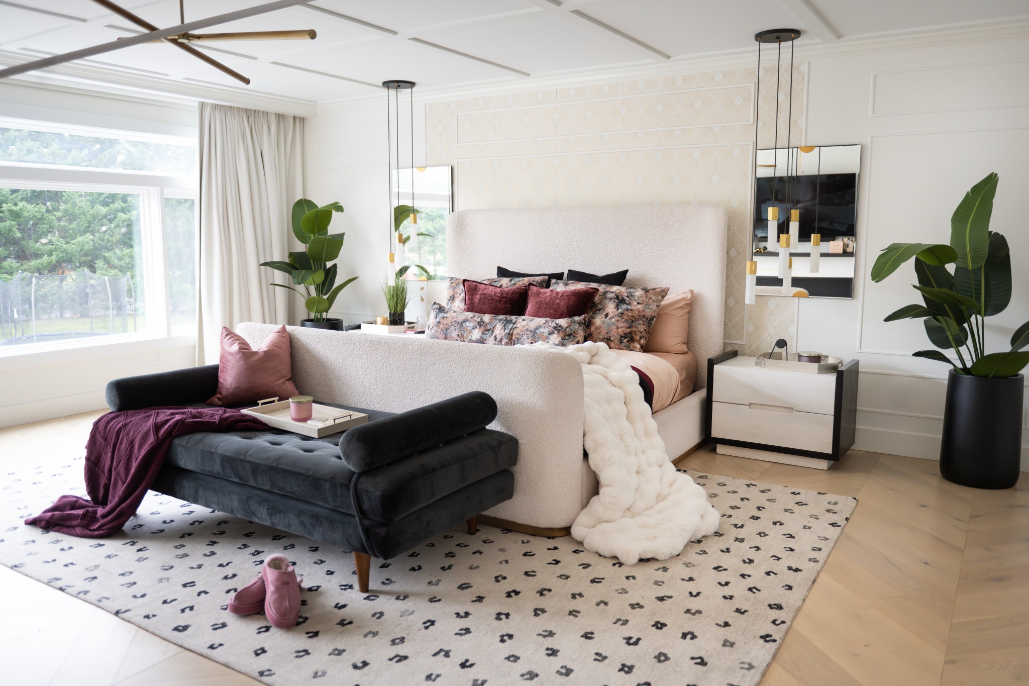 Living room with a large bed, black bench, and potted plants, bright window, and patterned rug. Word count: 20.