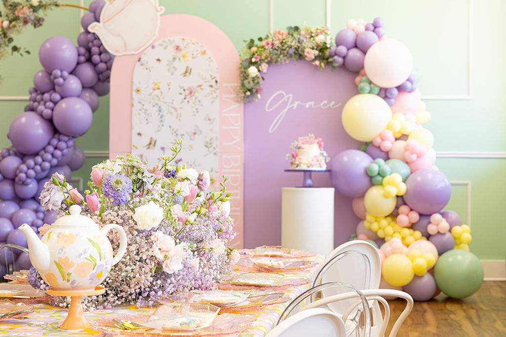 Decorative setup with purple, yellow, and white balloons, floral arrangements, a teapot, and a pink backdrop with writing.
