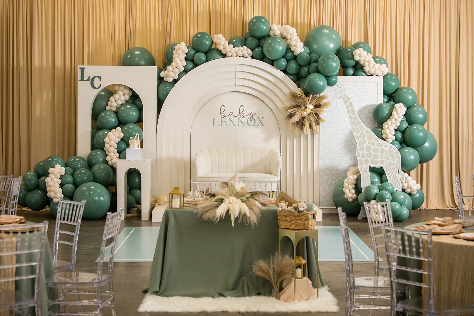 Decorative backdrop with green and white balloons, a white arch, and a table with a green cloth, set for a celebration.