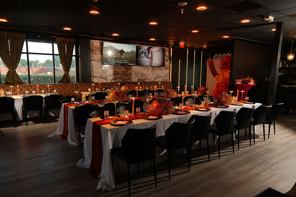 Long dining table set with white tablecloths, chairs, and decorations in a dimly lit restaurant with large windows and wall-mounted screens.