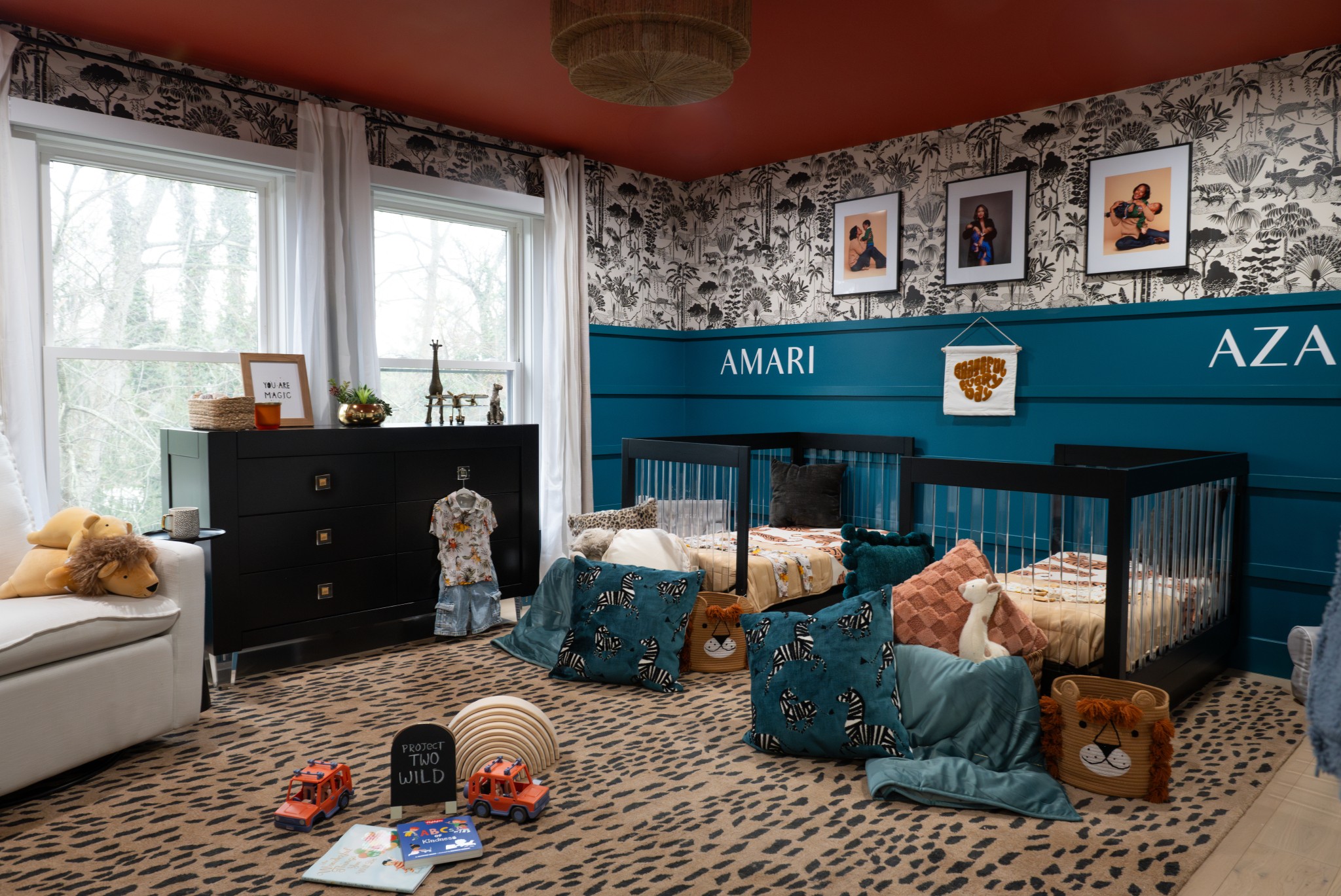 Children's bedroom with two cribs, a dresser, and toys on the floor, decorated with framed pictures and patterned wallpaper.