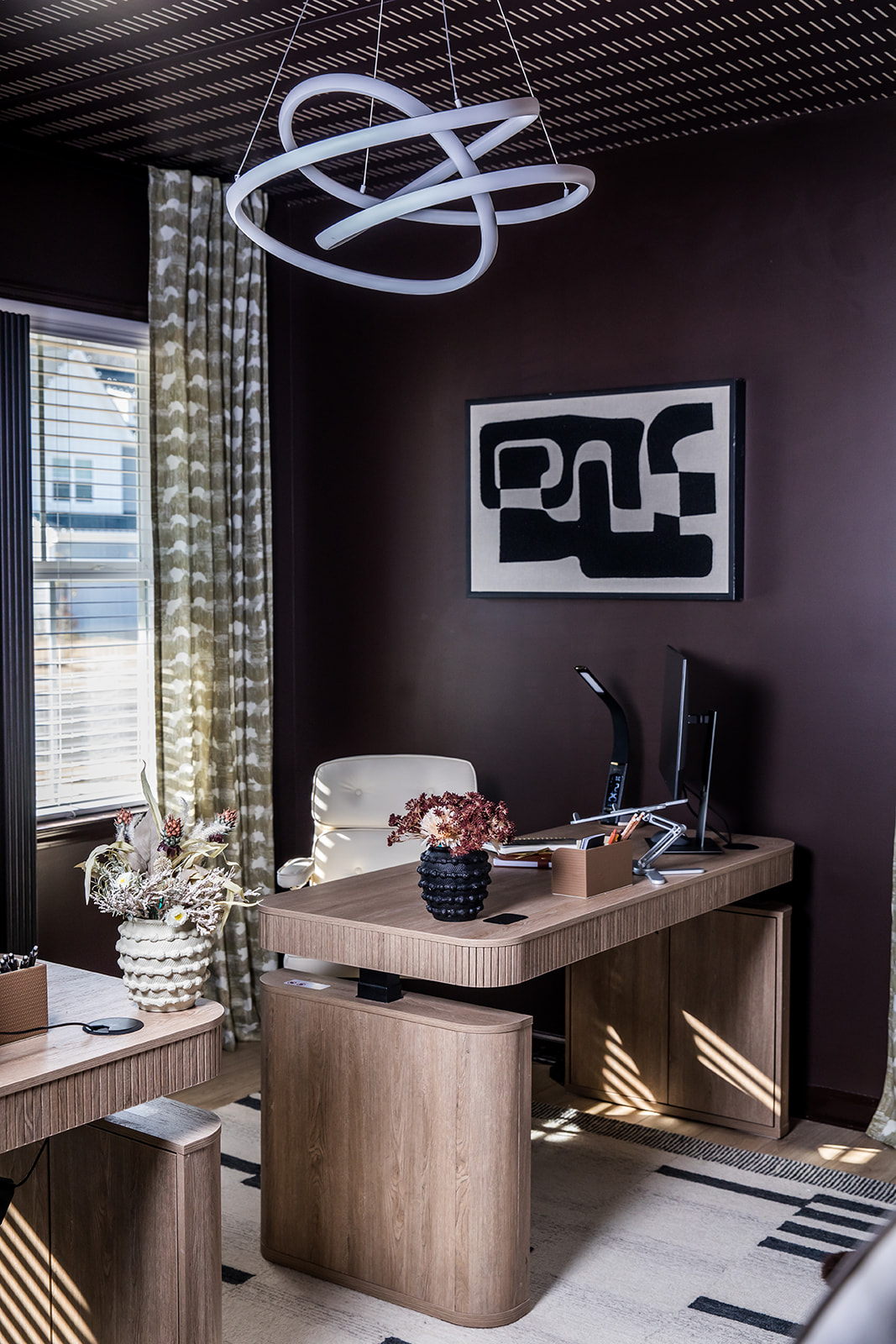 Office corner with desk, chair, artwork, and window with curtains, illuminated by natural light, modern ceiling light fixture.