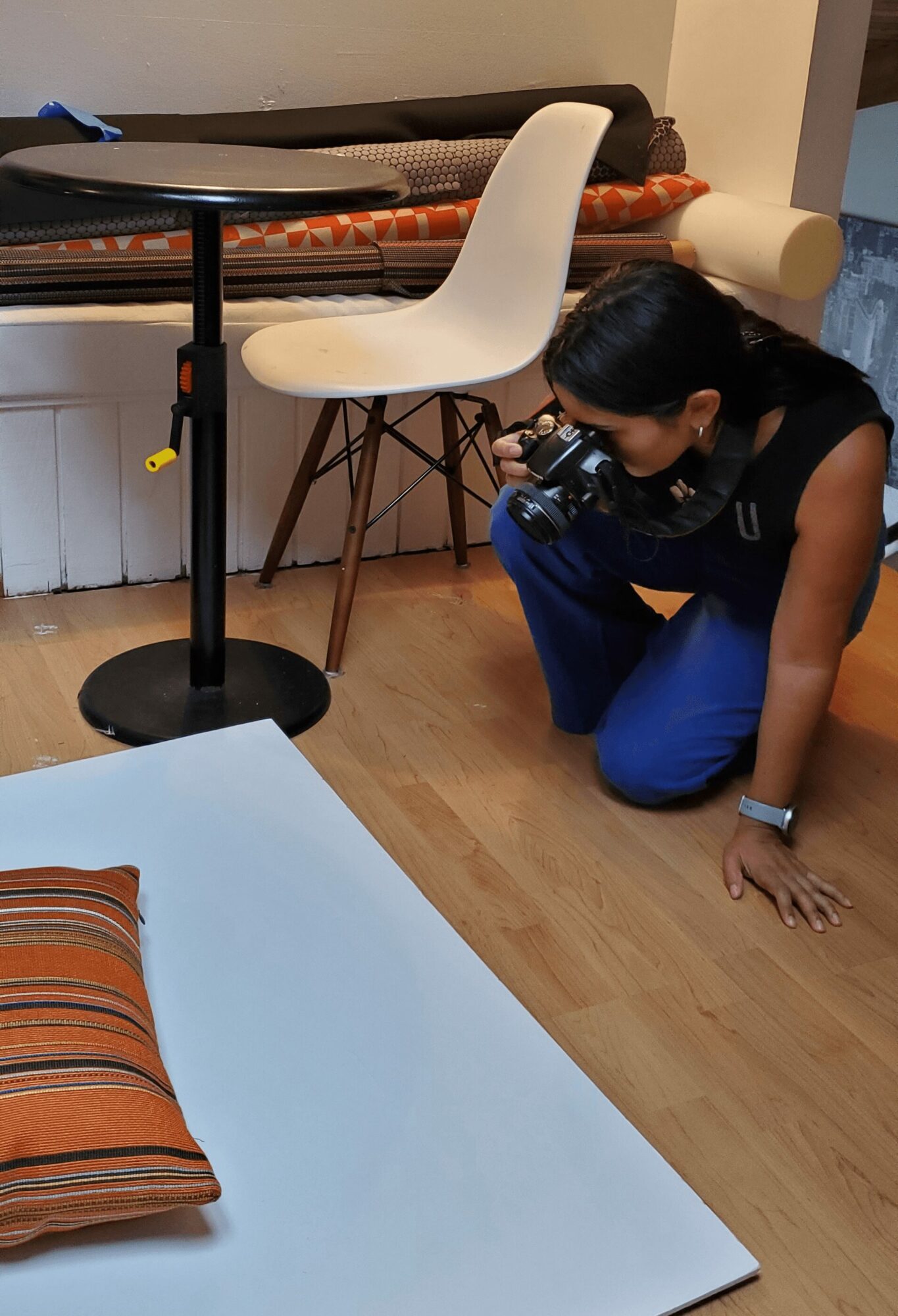 Person crouching on the floor taking a photo with a camera, near a white table and a pillow, in a room.
