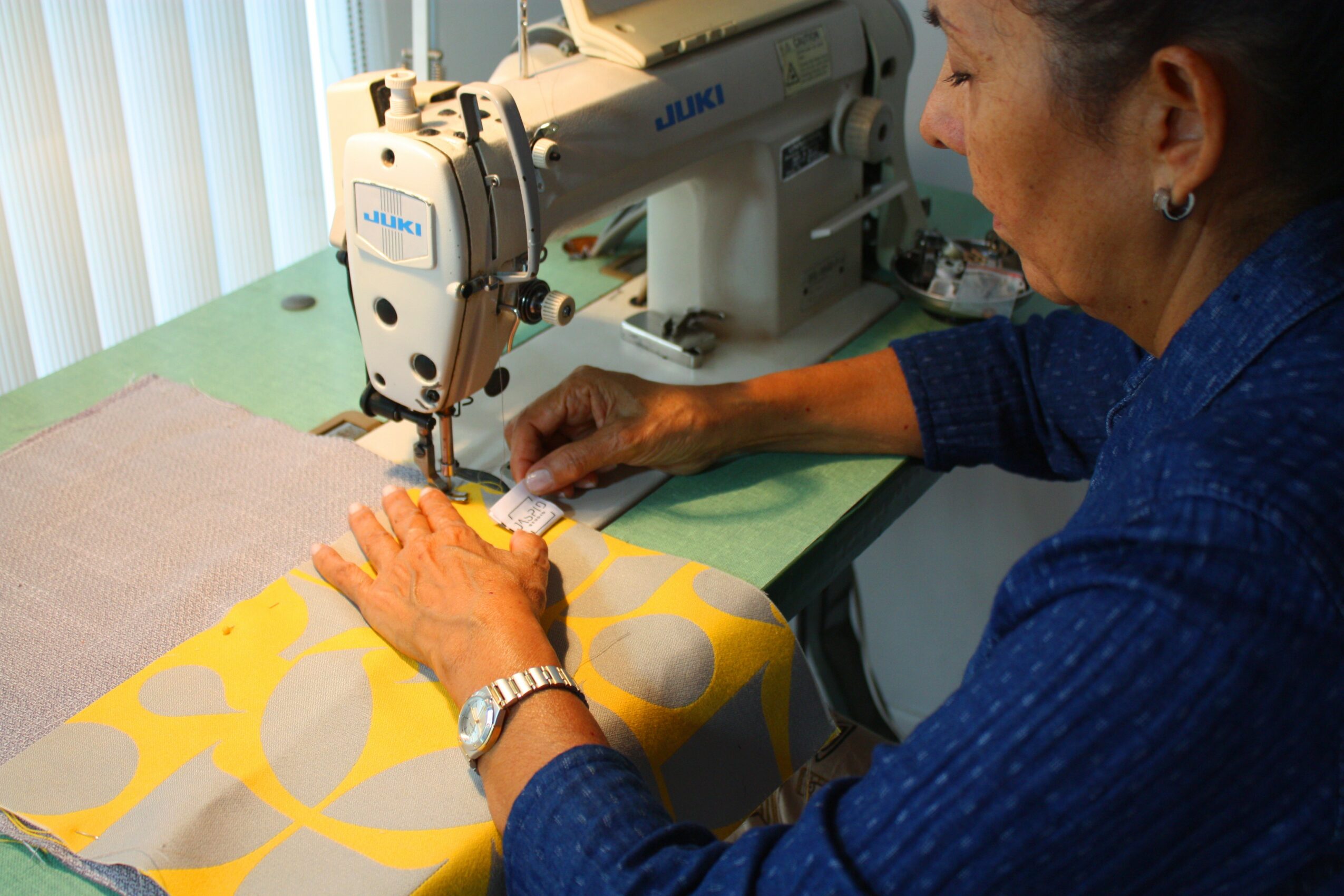 Woman sewing fabric with a sewing machine, wearing a blue shirt, working at a green table, with sunlight coming through blinds.