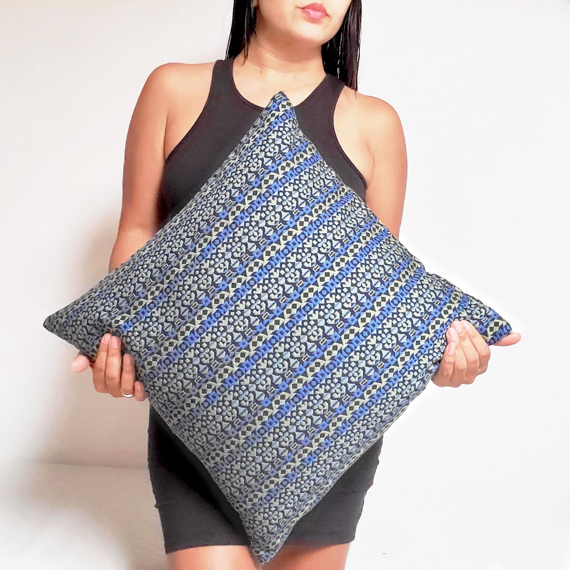 Person holding a patterned pillow with both hands, wearing a sleeveless black dress, standing against a plain background.
