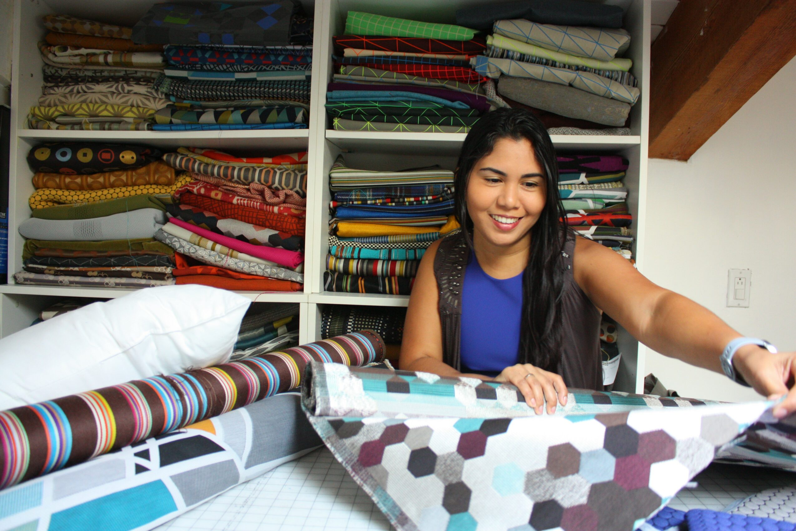 Woman smiling at fabric store with shelves of colorful fabric behind her, reaching for fabric on table.