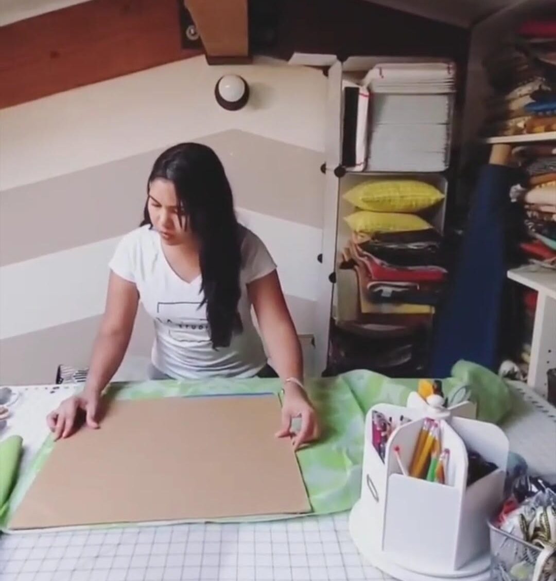 Woman standing at a table with craft supplies, working on a large piece of paper or cardboard, in a room with shelves.