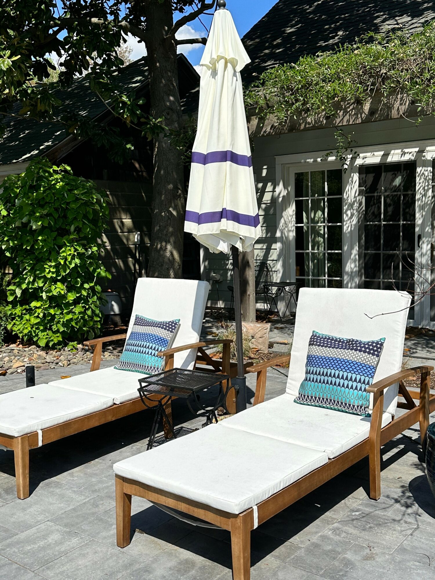 Two white lounge chairs with blue patterned pillows, a closed white umbrella, and a garden with greenery and a building in the background.