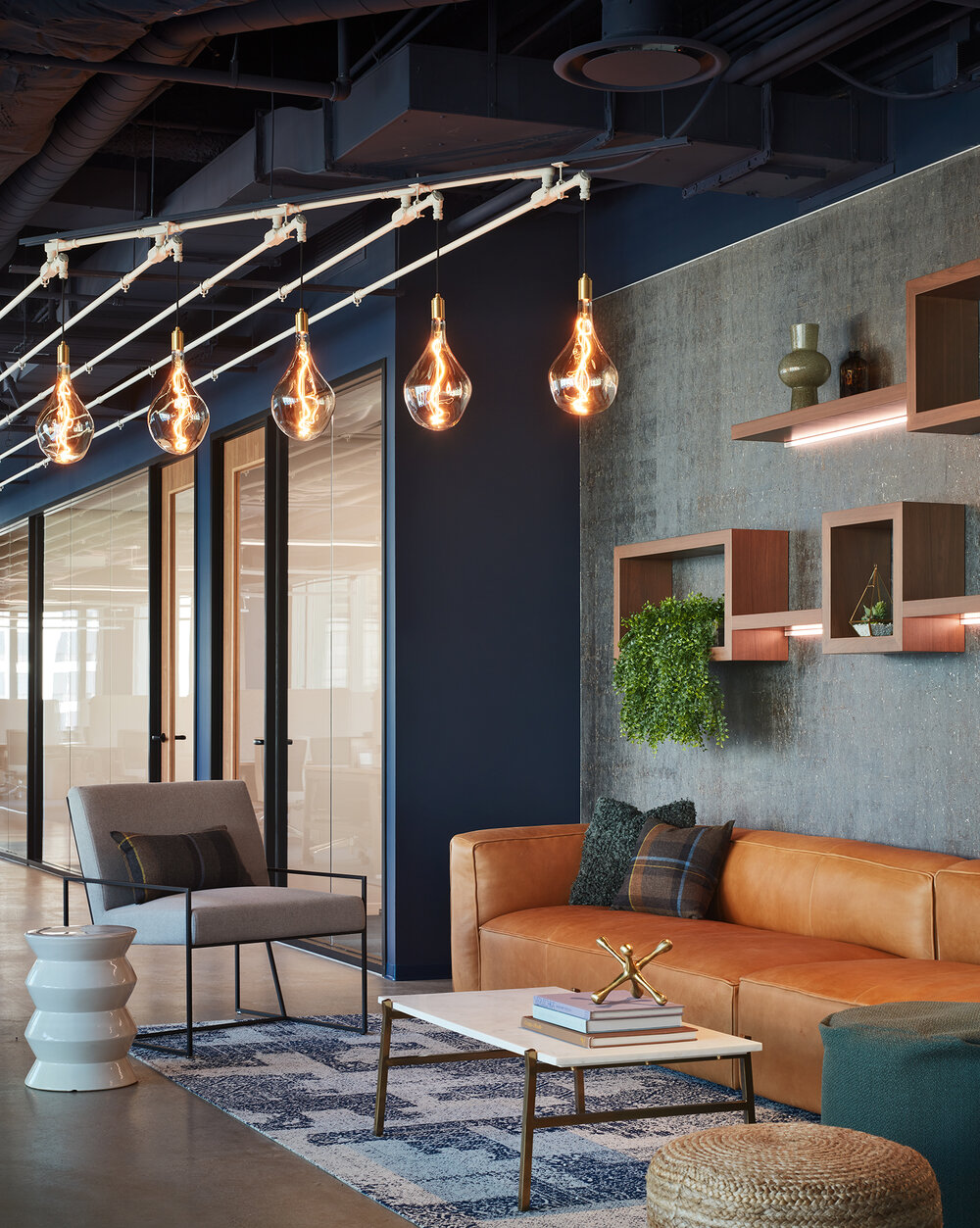Modern living room with a brown leather sofa, wall shelves, and hanging pendant lights, featuring a large window and decorative items.