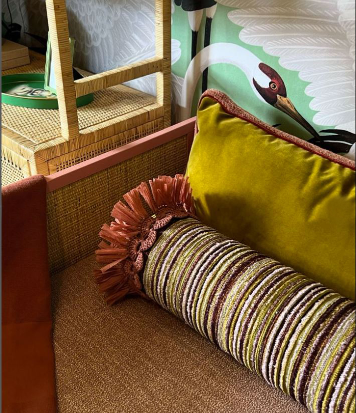 Close-up of a sofa with a green pillow and a striped cylindrical cushion, with a woven chair and decorative wall in background.