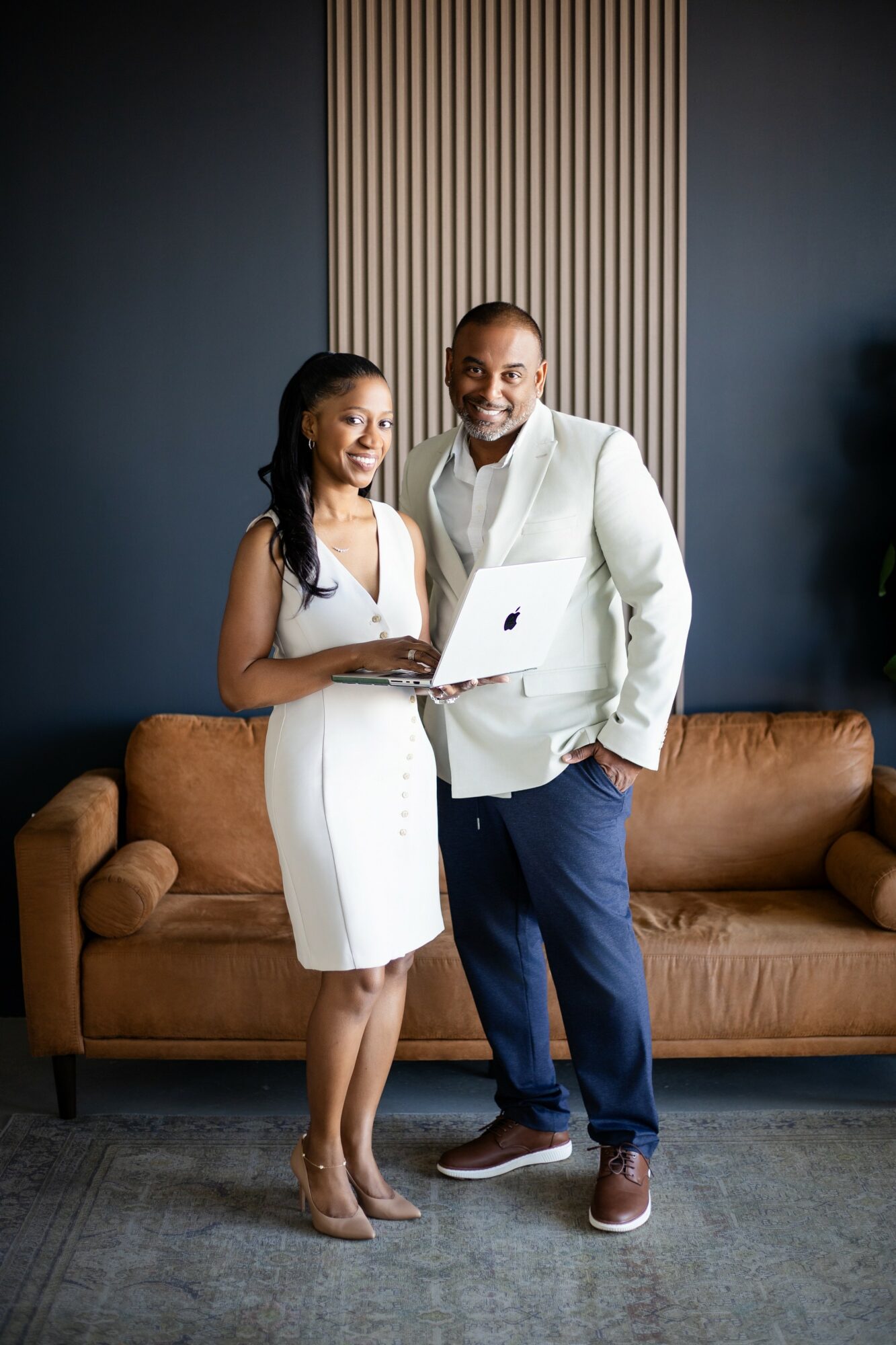 A man and woman stand together in a modern room, smiling, with a laptop in the woman's hands, in front of a brown sofa.