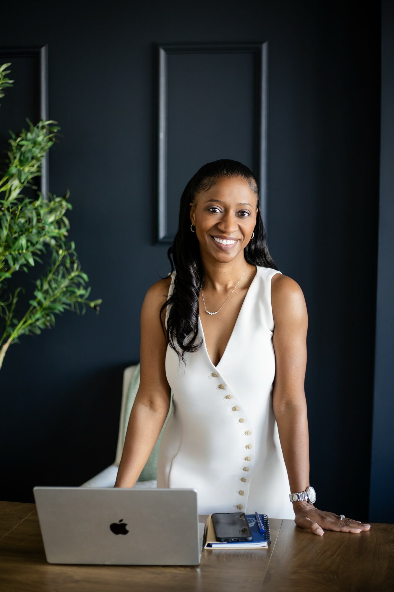 Smiling woman in white dress standing behind a desk with a laptop and smartphone, dark wall background.