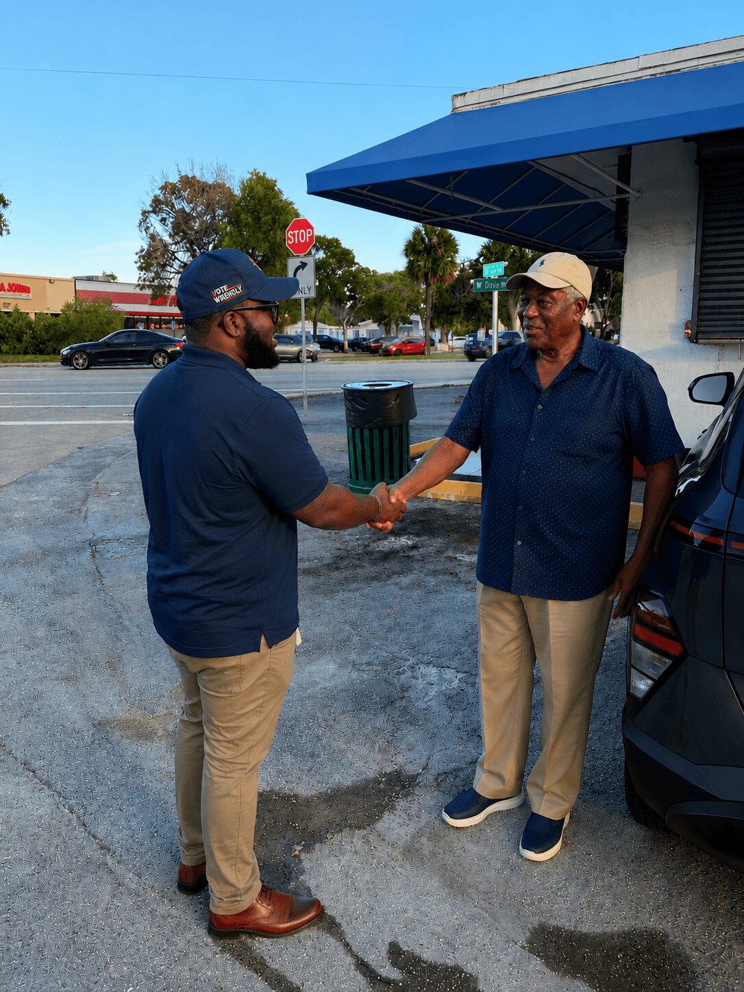 Two men shake hands outside a building, one wearing a cap and glasses, the other in a beige hat and glasses.