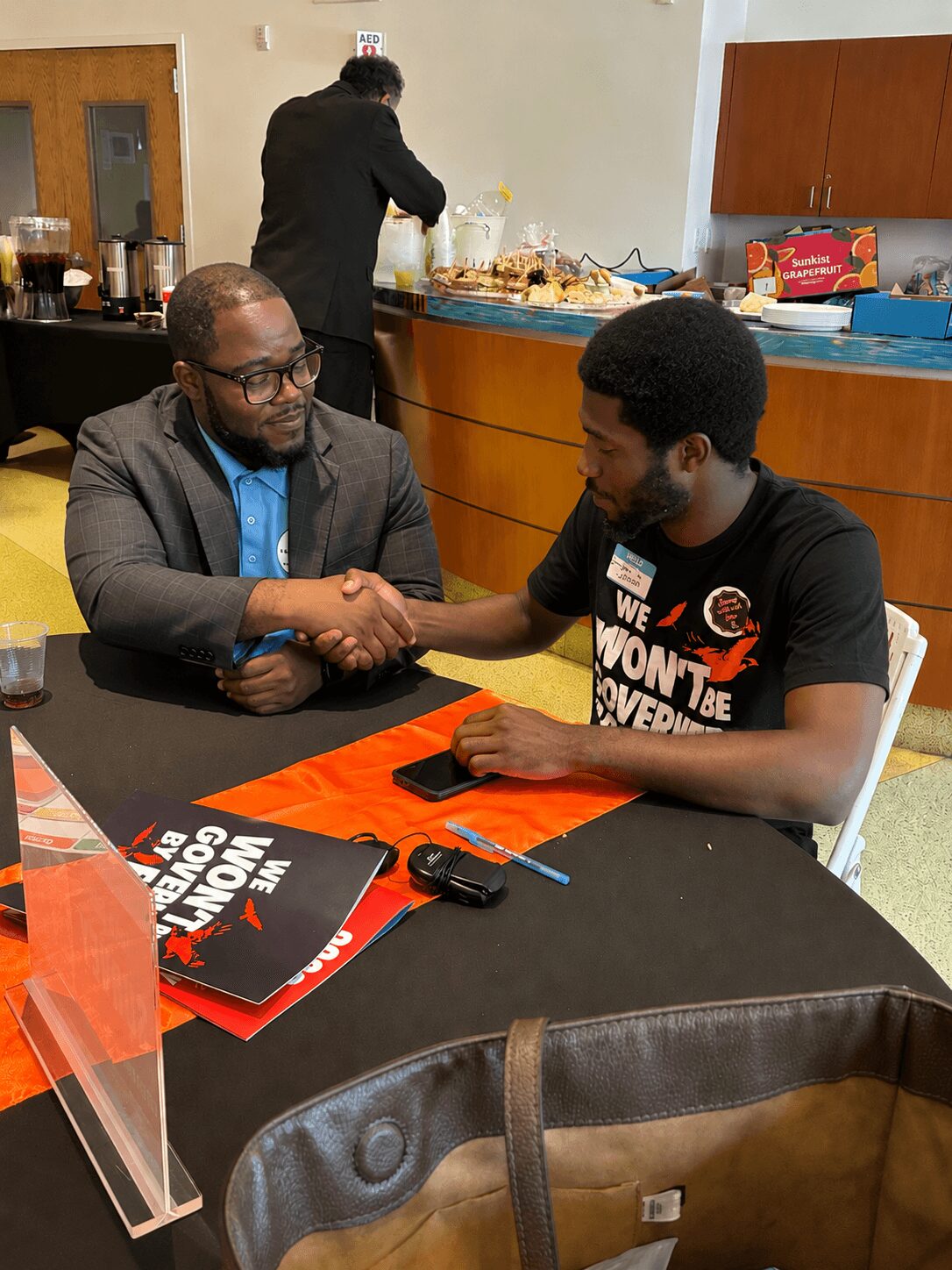 Two men shaking hands at a table, one wearing glasses and a suit, the other in a black T-shirt, in a room with a buffet in the background.