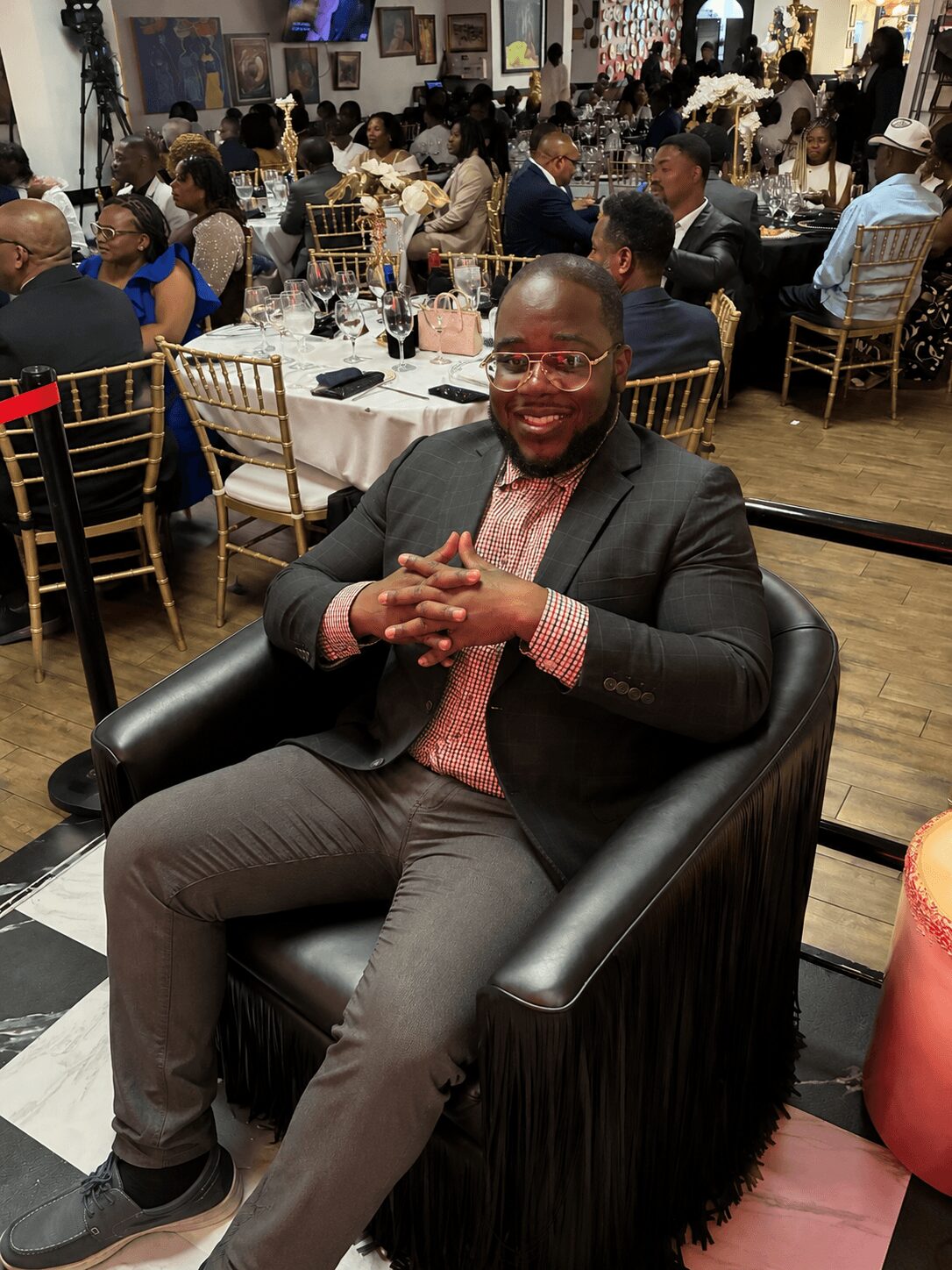 Man sitting in a black armchair at a formal event, smiling, with a large crowd in the background.