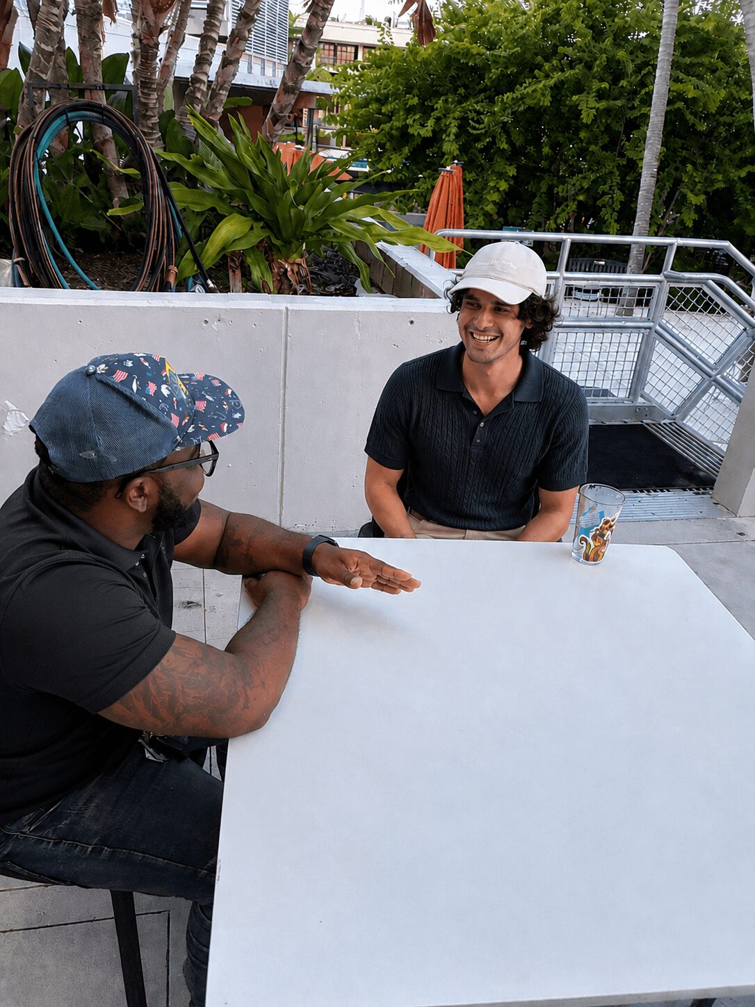 Two people sitting at a white table outdoors, smiling and talking, with trees and buildings in the background.
