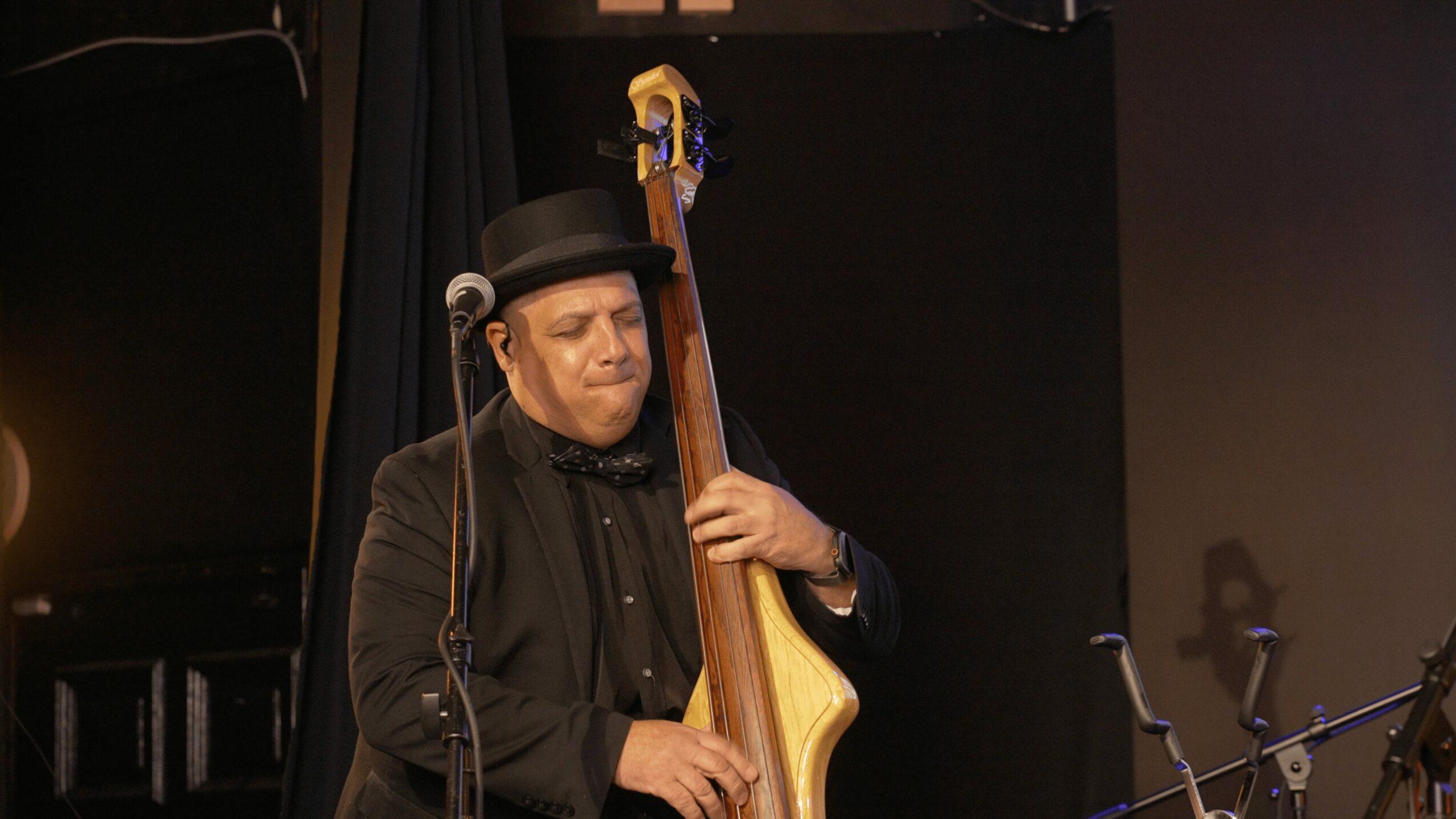 Musician playing a double bass on stage, wearing a black hat and dark clothing.