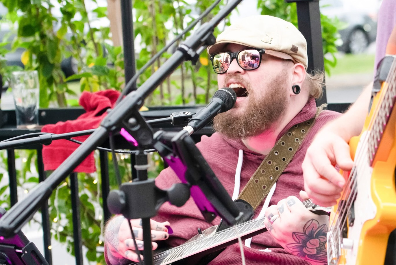 Man with glasses and a cap singing into a microphone while playing guitar outdoors, surrounded by greenery.
