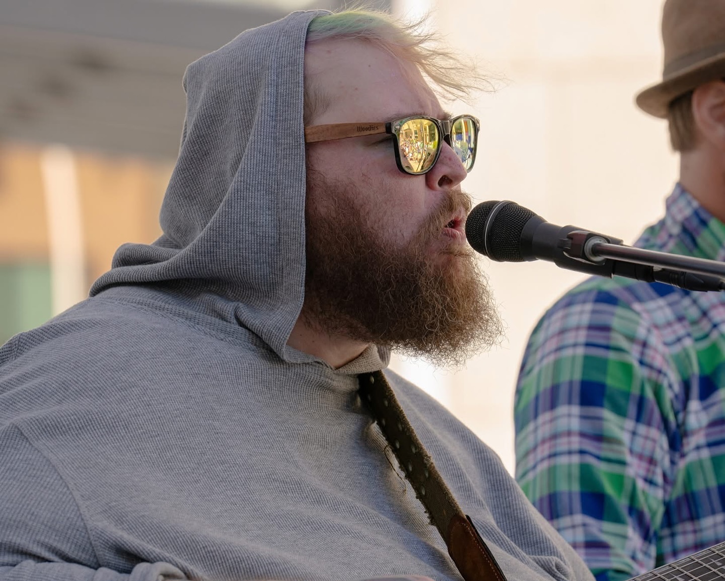 Bearded man wearing sunglasses and a gray hoodie singing into a microphone outdoors.