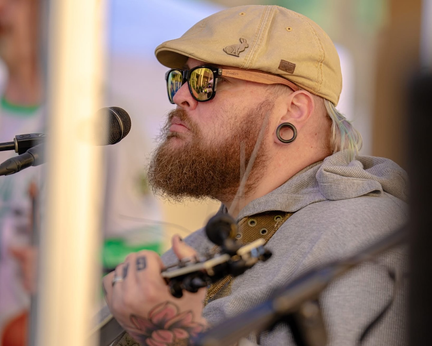 Man with a beard, sunglasses, and a cap playing guitar near a microphone, outdoors, with blurred background.