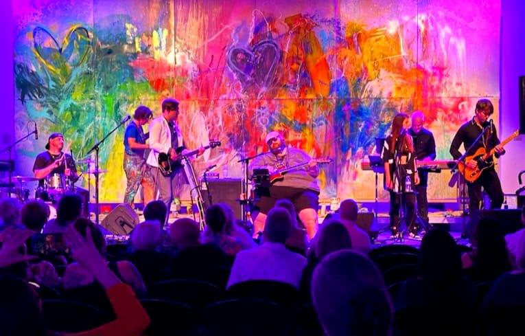 Band performing on stage with colorful abstract background, audience watching, some taking photos, vibrant atmosphere.