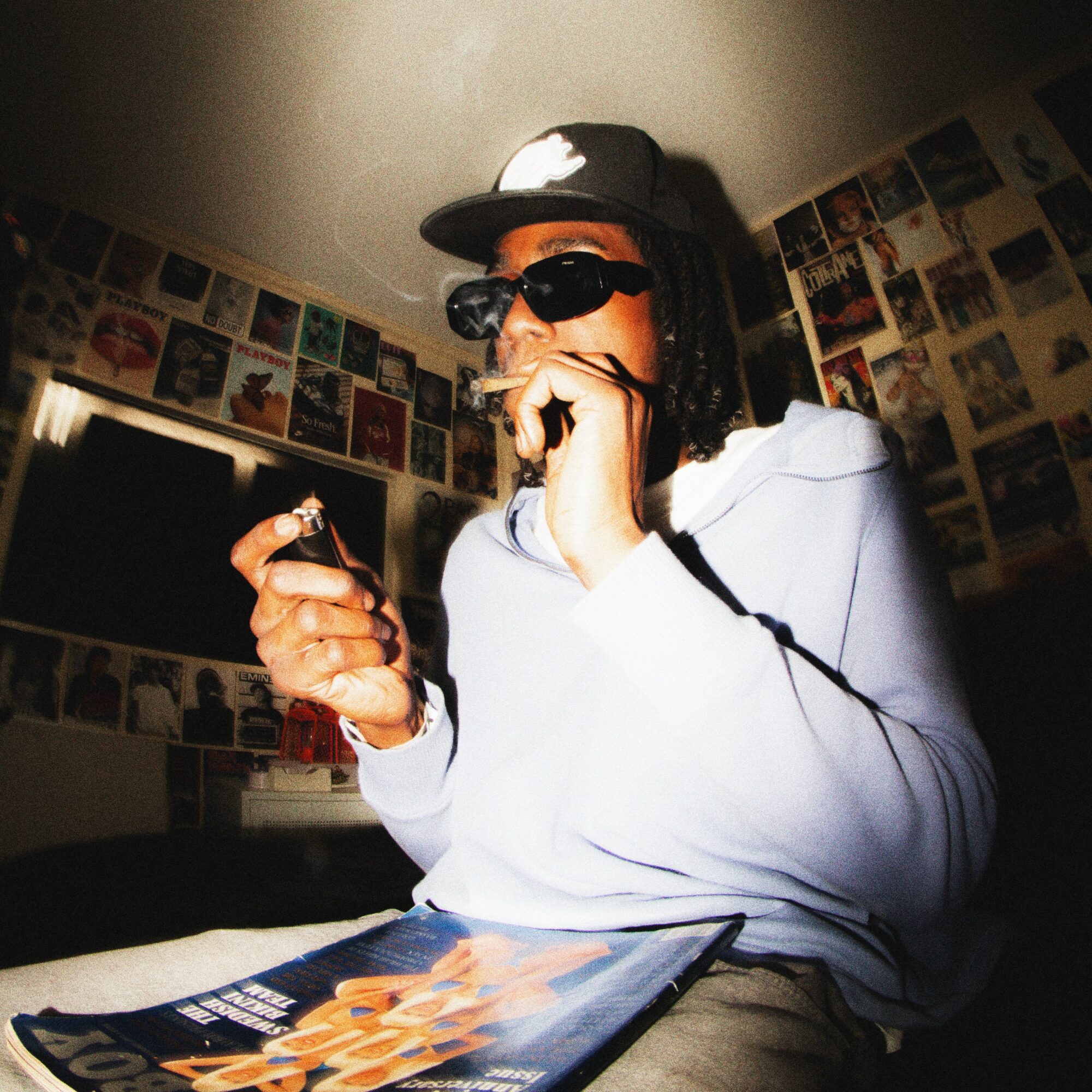 Person wearing sunglasses, a cap, and hoodie, holding a lighter and a phone, in a room with wall posters.