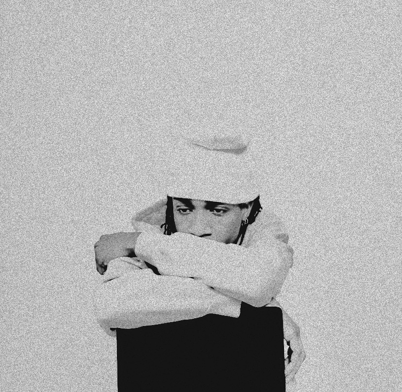 Person with arms crossed, wearing a hat, looking to the side, in black and white, plain background.