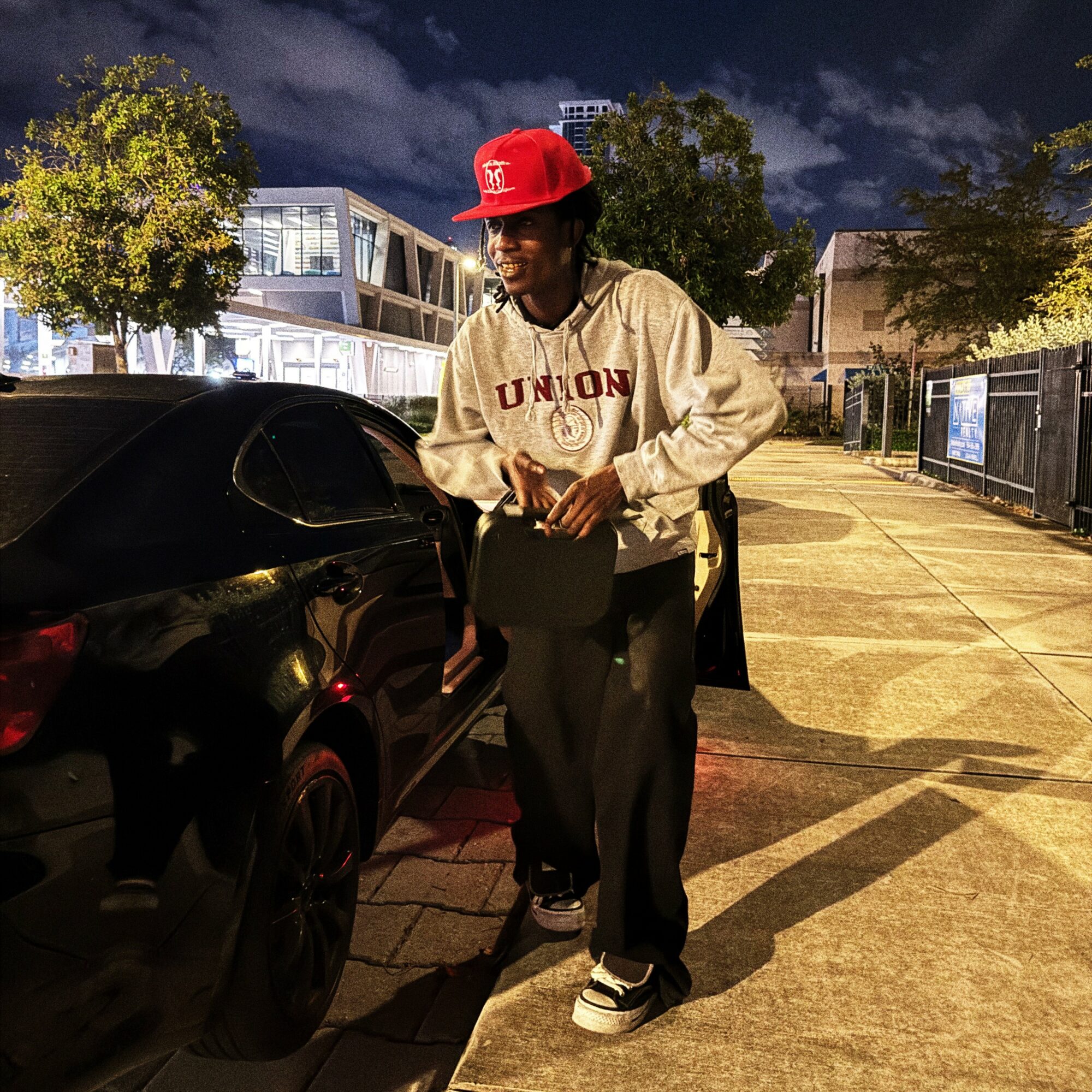 Person walking on sidewalk at night, wearing a red cap, beige jacket, and black pants, holding a bag, with trees and buildings in background.