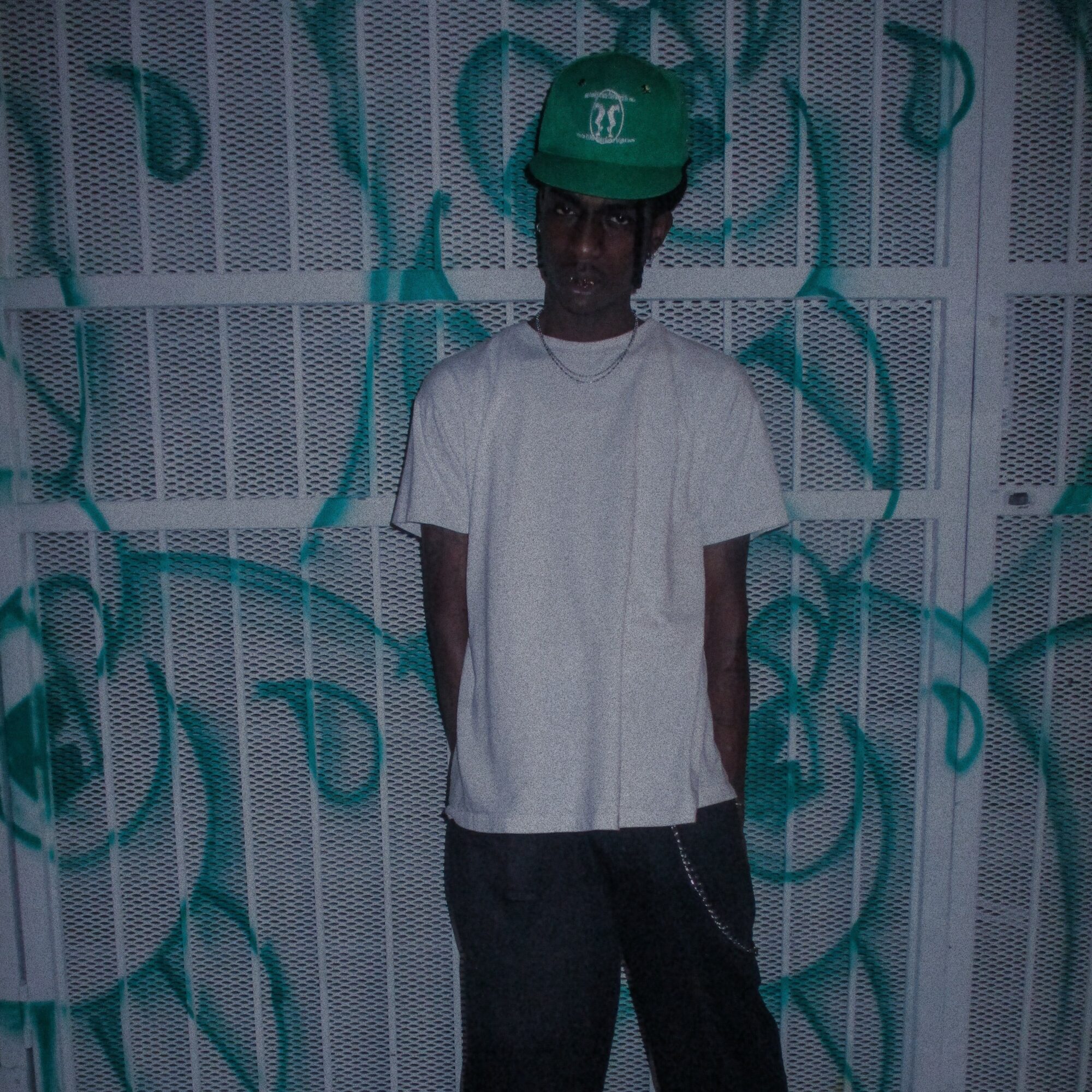 Person wearing a green helmet and white shirt standing against a wall with blue graffiti, facing forward.