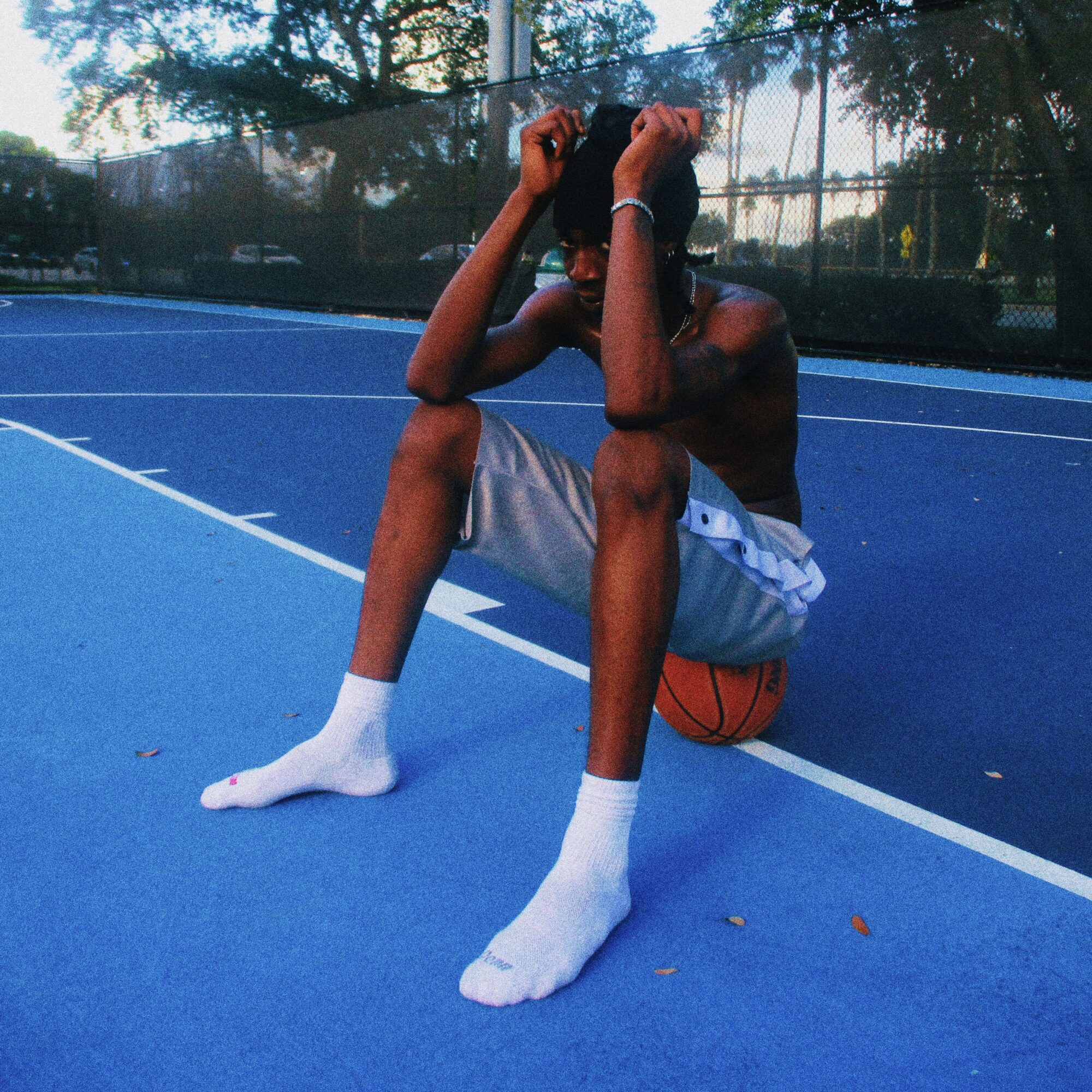 Young person sitting on a basketball court with legs spread, holding their head, wearing white socks and shorts, with a basketball nearby.