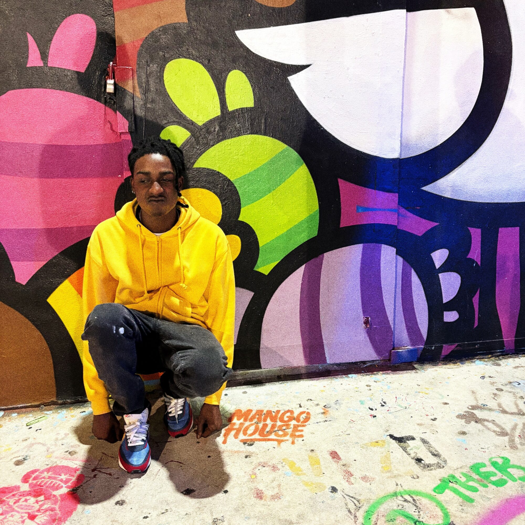 Person crouching in front of a colorful graffiti wall, wearing a yellow hoodie and gray pants.