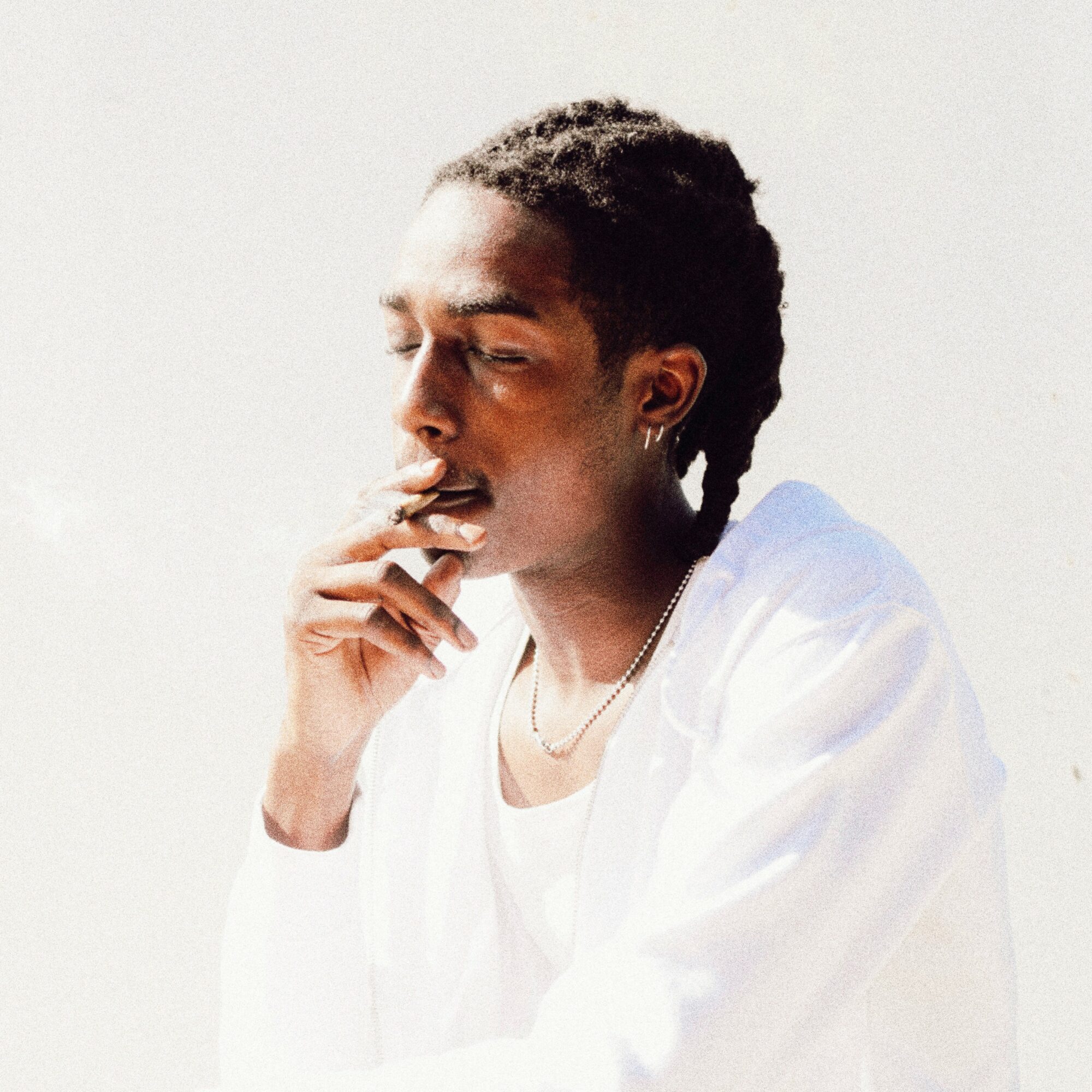 Person with short curly hair and earrings, wearing a white shirt, smoking against a plain background.