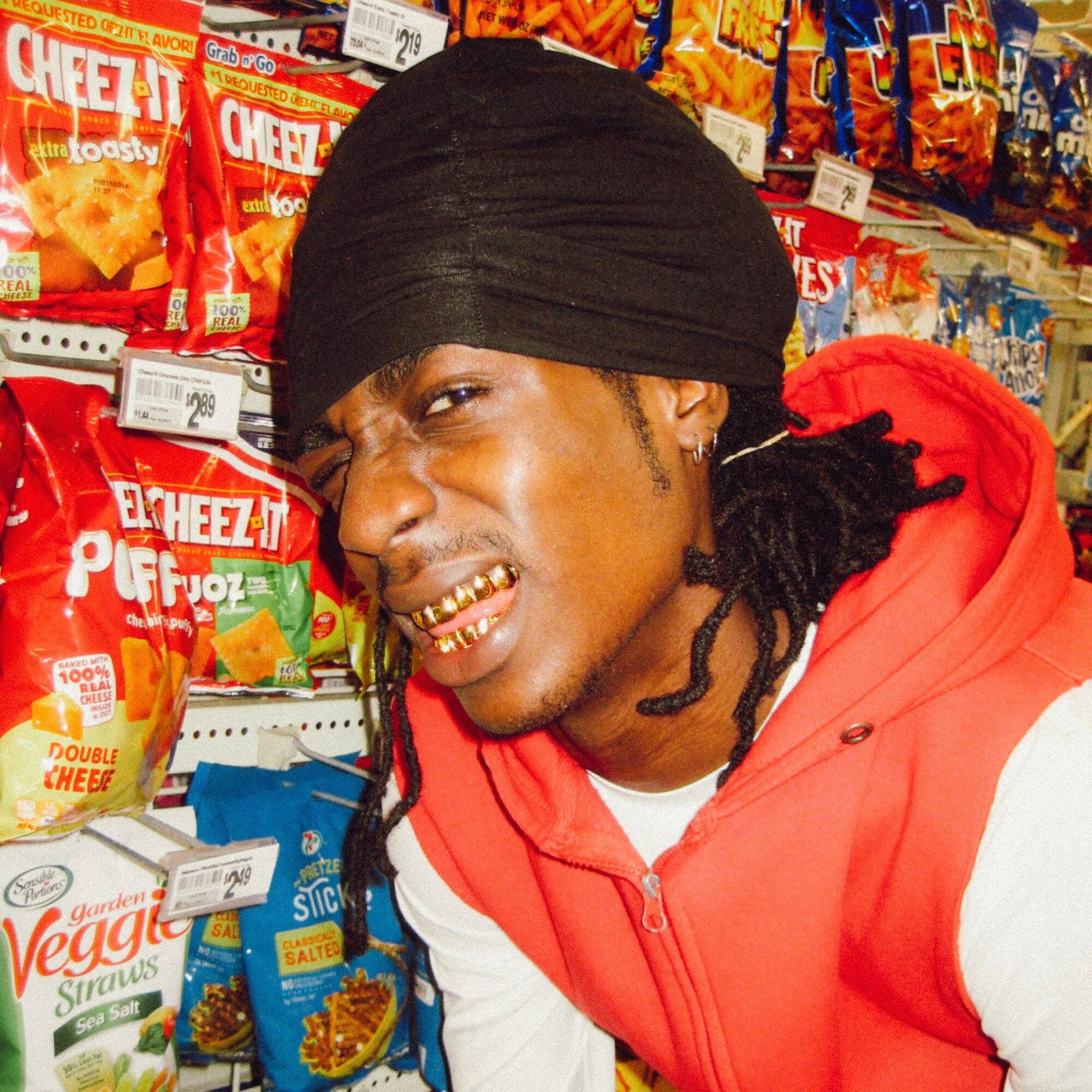 Young man with a black headwrap, gold grill, smiling in a grocery store aisle with snack bags behind him.