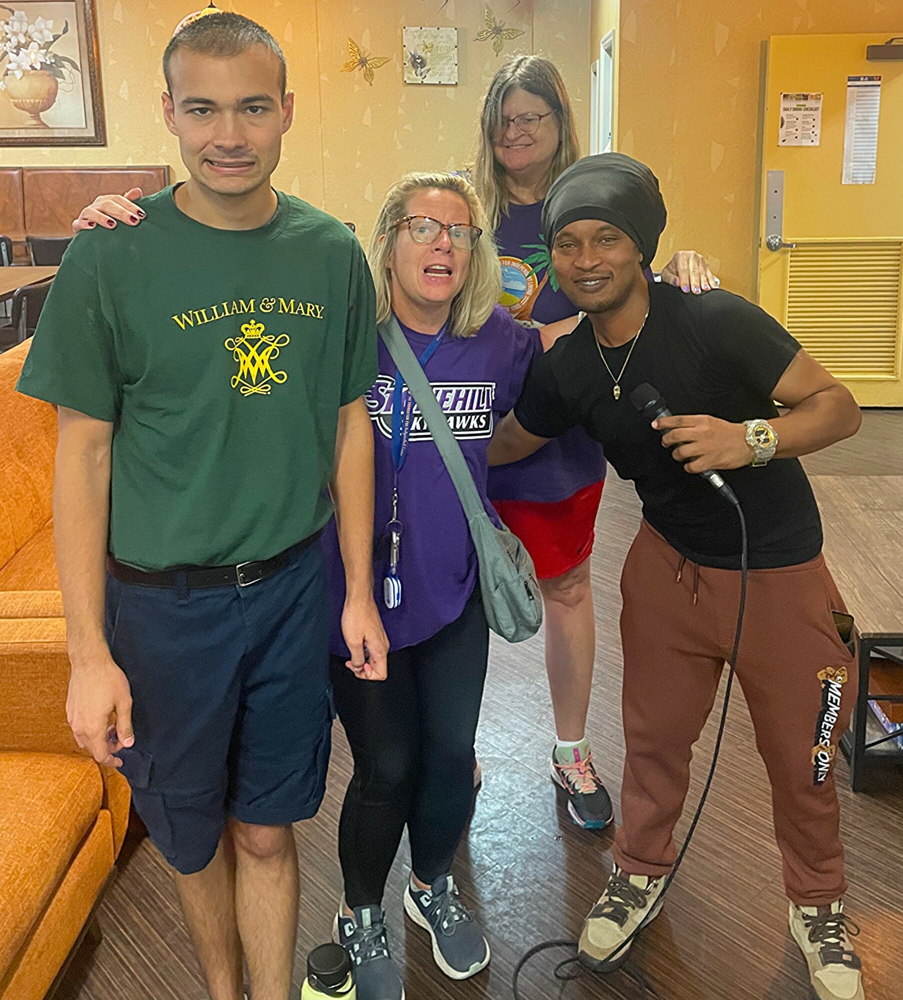 Four people standing indoors, smiling, with a yellow wall and furniture in the background.