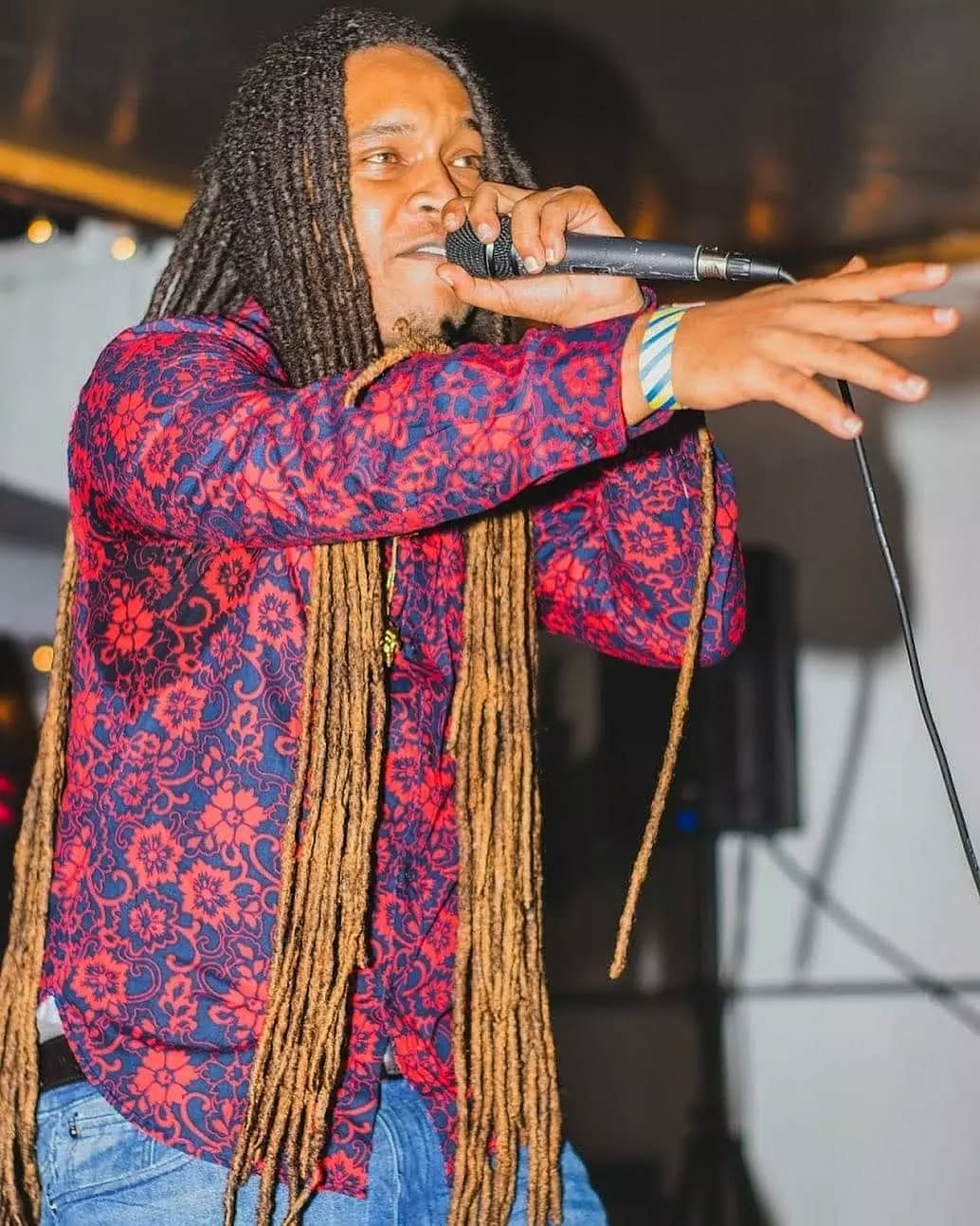 Person with long dreadlocks singing into a microphone, wearing a colorful patterned jacket, gesturing with their hand.