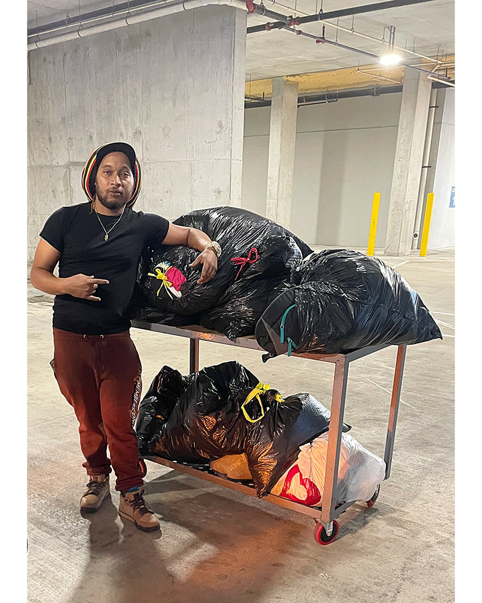 Person standing next to a cart with large black bags in an indoor parking garage.
