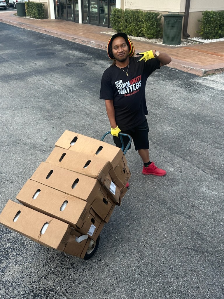 Person standing on street with a cart of cardboard boxes, wearing black shirt, yellow gloves, and red shoes.