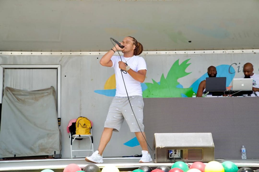 A woman singing into a microphone on stage with two people working at a table behind her, colorful balloons in front.